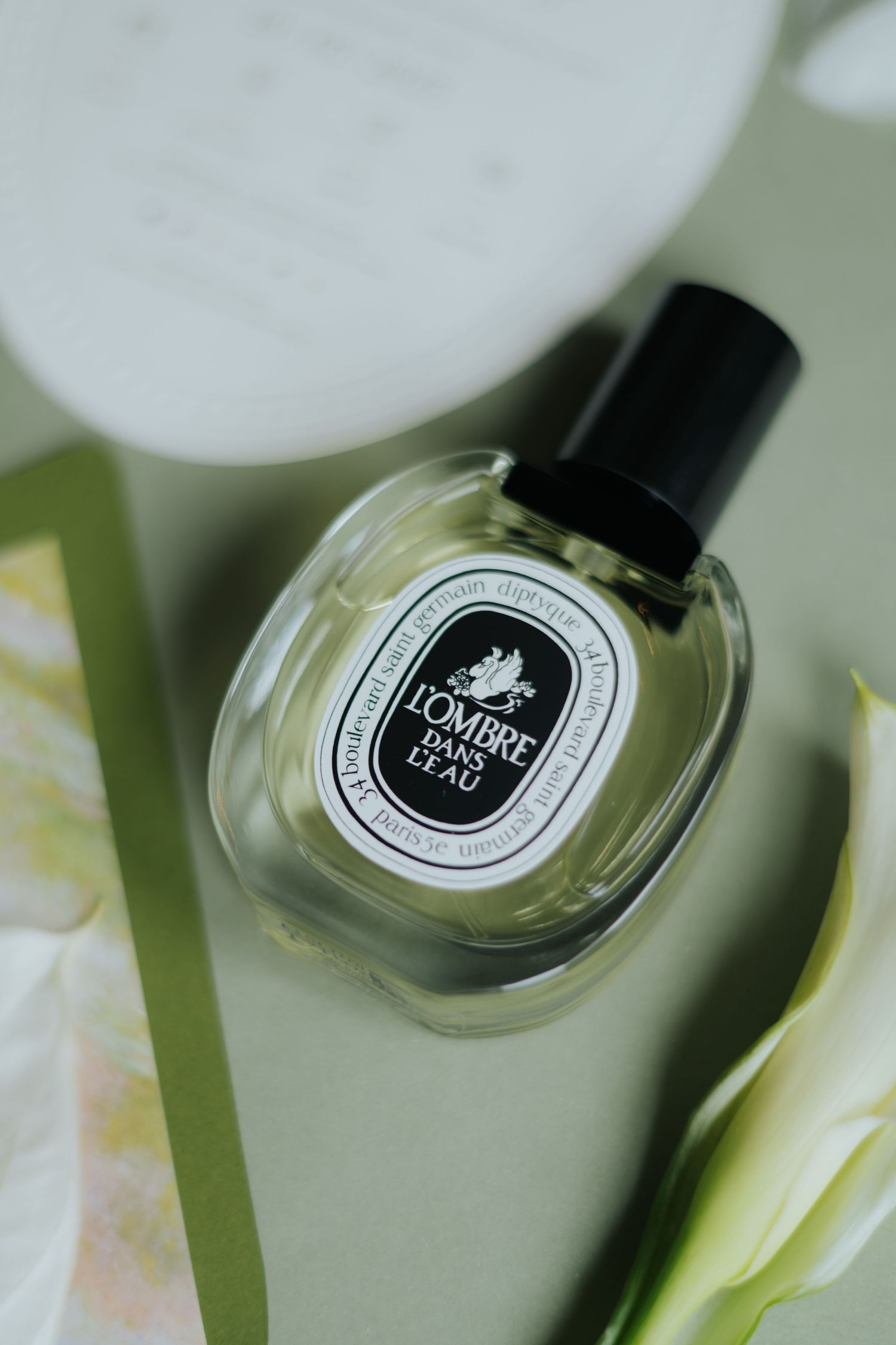 Diptyque L'Ombre Dans L'Eau eau de parfum bottle resting on a green surface with white flowers.