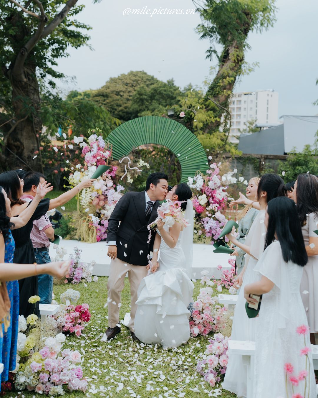 Couple kissing at outdoor wedding ceremony with floral arch, green fan backdrop, and guests celebrating with confetti.