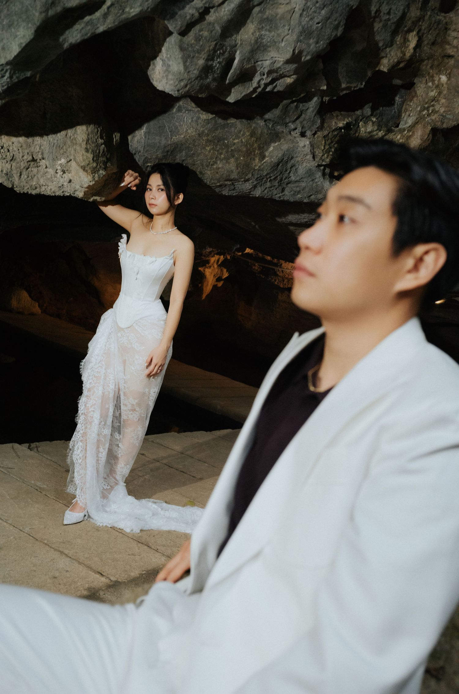 Couple in wedding attire pose near rocky cave, woman in white lace gown, man in white suit in foreground.