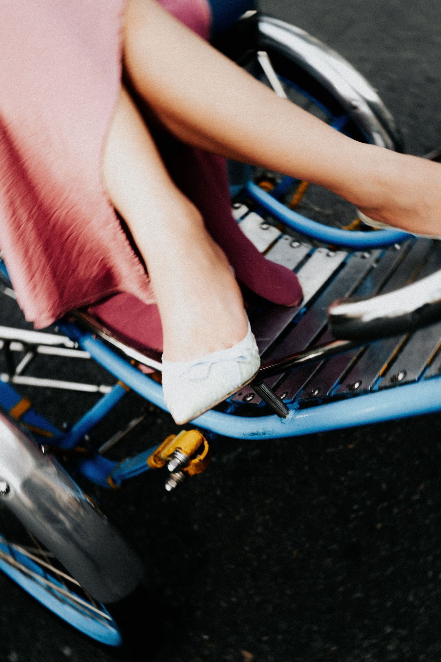 Close-up of a woman's legs in white flats and pink skirt sitting on a blue bicycle rickshaw on pavement.