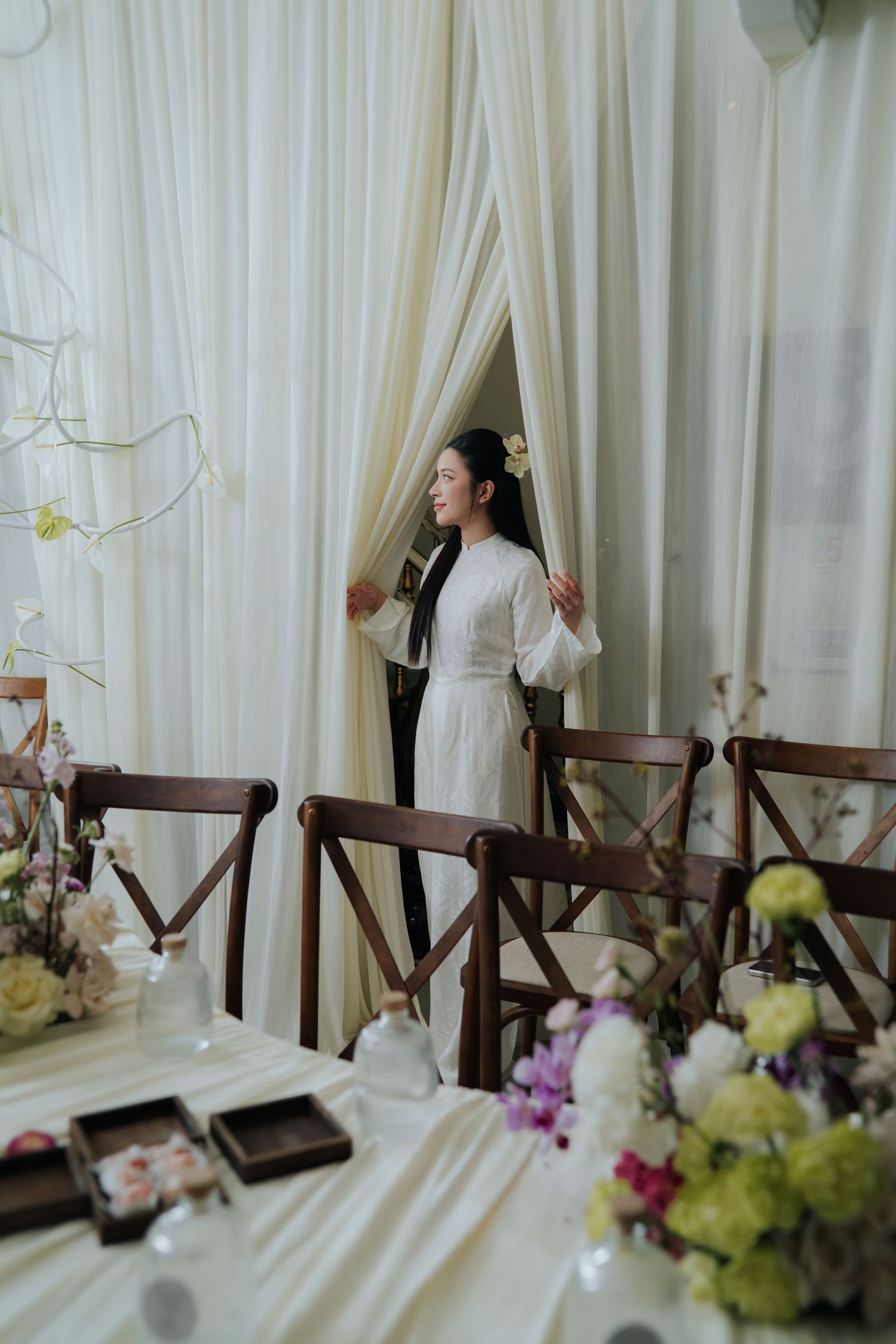 Elegant woman in white dress standing between draped curtains at a floral-decorated wedding reception table.