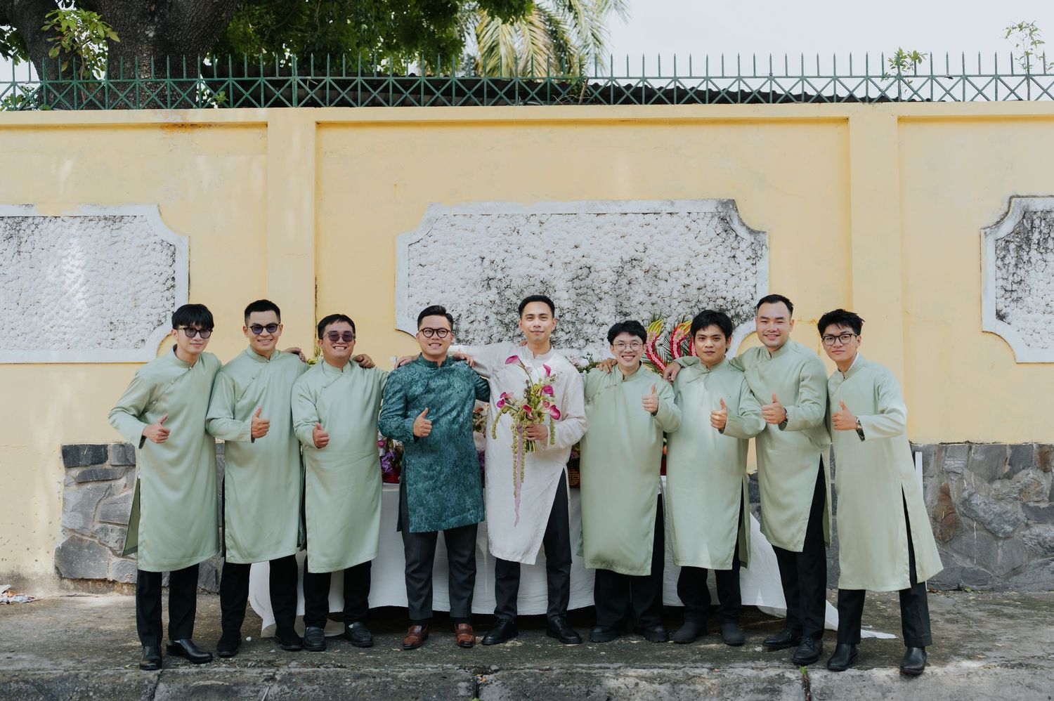 Group of men in matching light green traditional Vietnamese ao dai attire posing together outdoors near a yellow wall.