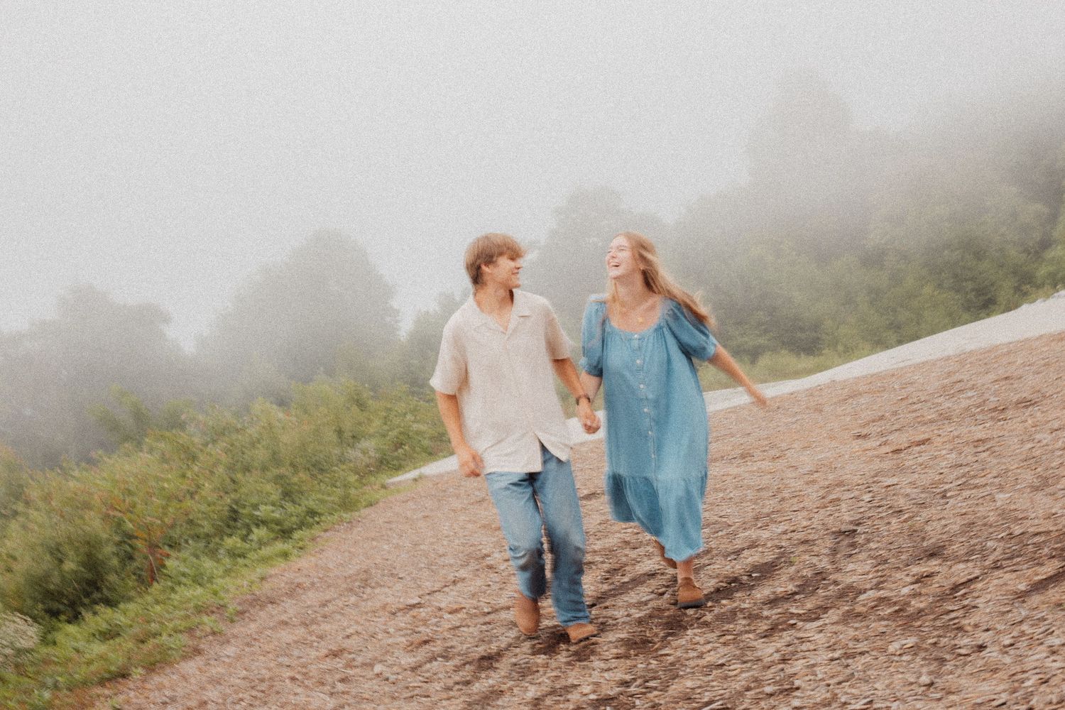 Young couple holding hands walking on foggy mountain path surrounded by lush green vegetation.