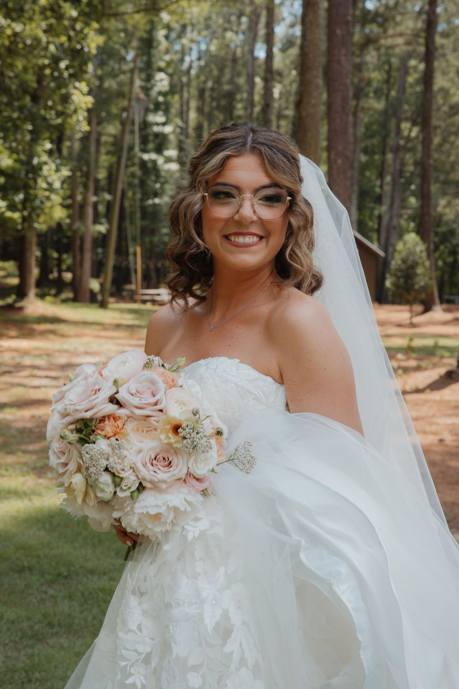 Smiling bride wearing glasses and veil holds blush pink rose bouquet outdoors among pine trees.
