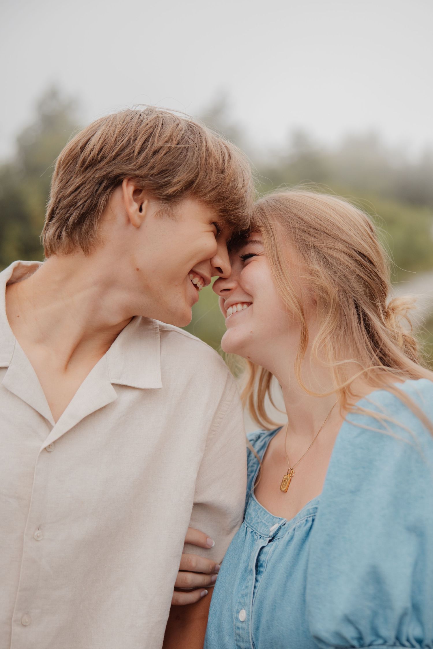 A young couple smiles and touches foreheads in a romantic outdoor engagement photo on a misty day.