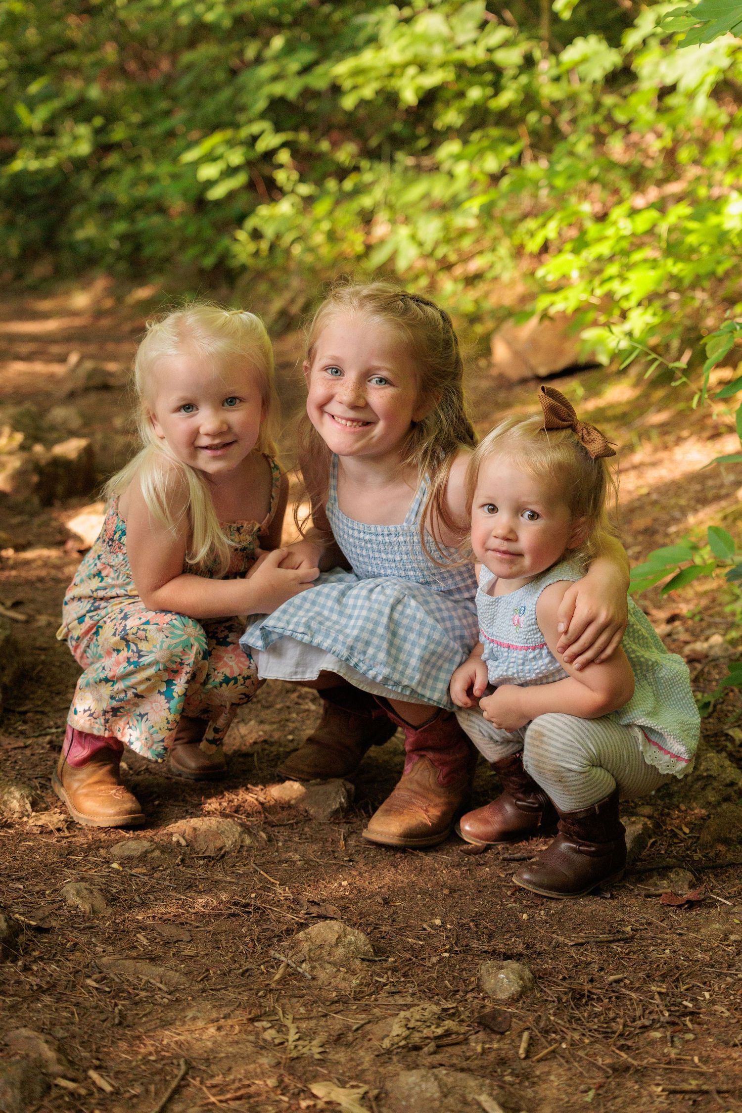 Three young blonde girls in dresses crouch together smiling in a sunlit forest setting.