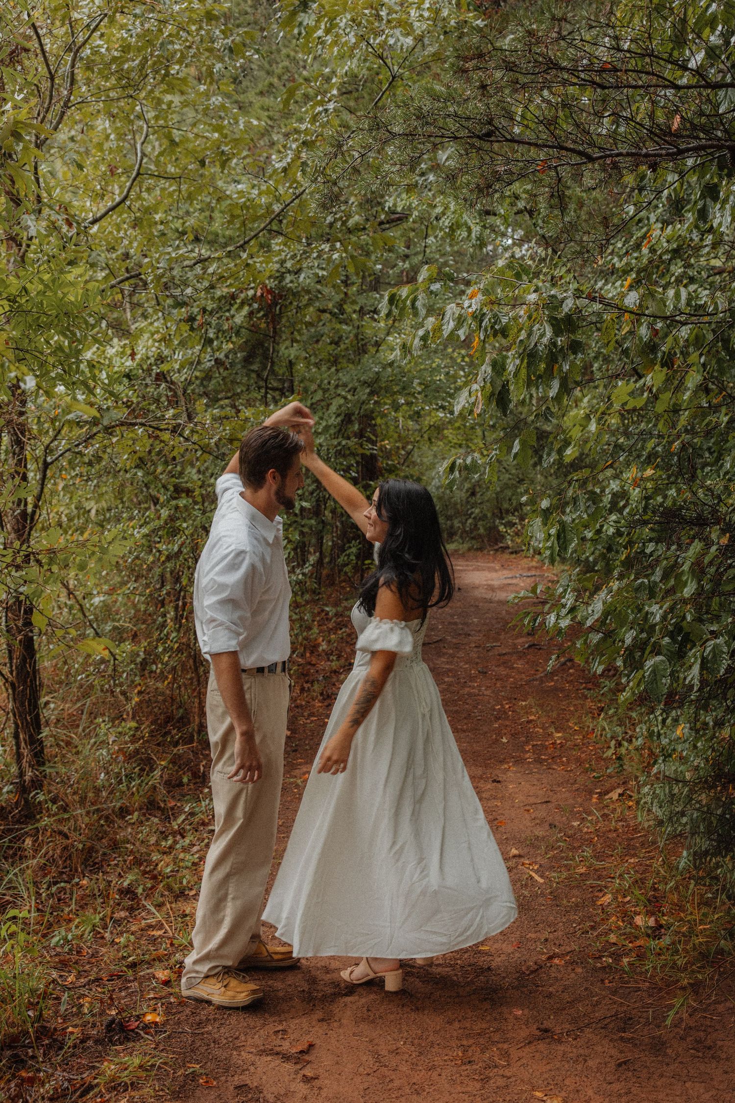 Couple dancing on a dirt path in a lush green forest, woman in a white dress being twirled.