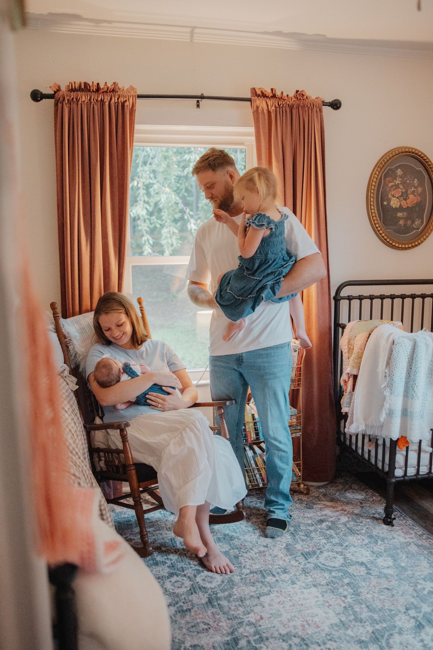Family with newborn baby and toddler in a cozy nursery with pink curtains and vintage decor.