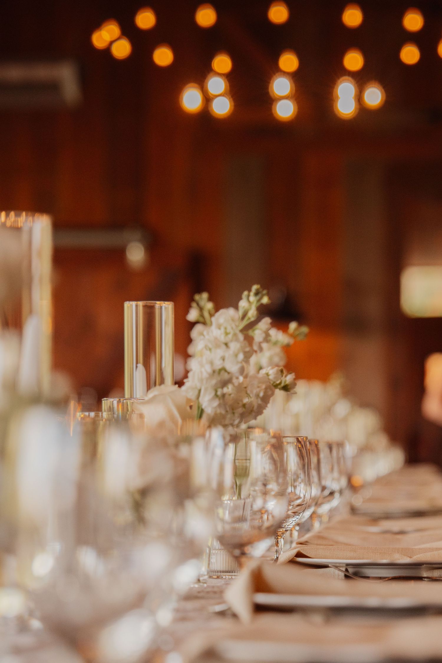 Elegant wedding reception table with white floral centerpieces, crystal glassware, and warm bokeh string lights.