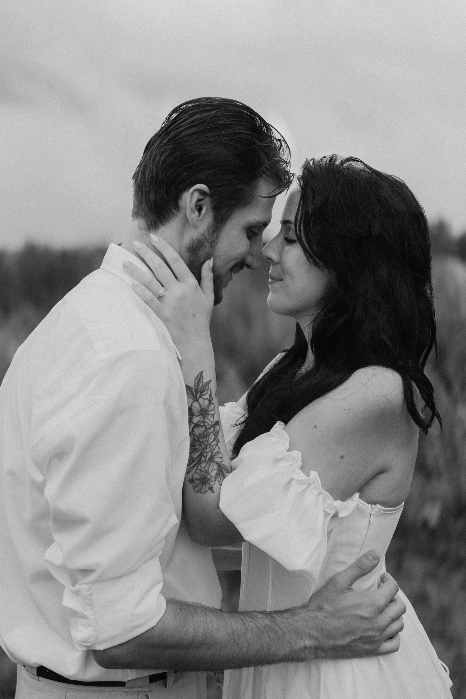Black and white photo of couple nearly kissing, woman in off-shoulder dress with man's floral arm tattoo.