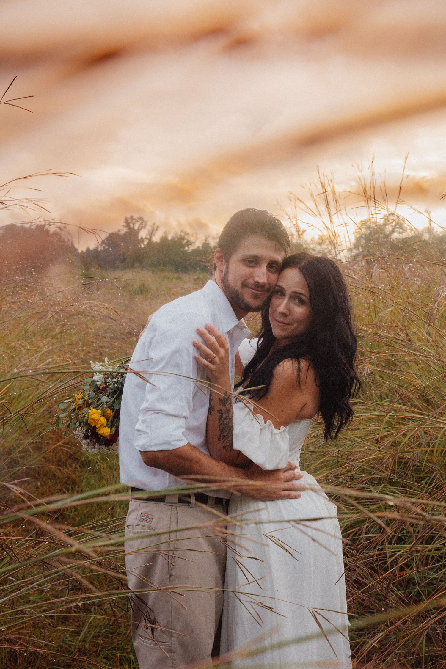 Couple embracing in tall golden grass at sunset holding a wildflower bouquet.