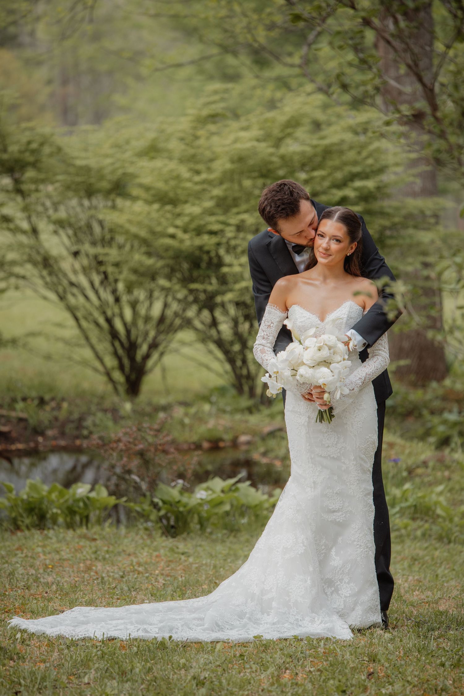 Groom in black tuxedo embraces bride in lace mermaid gown holding white bouquet, posing outdoors in lush green garden.