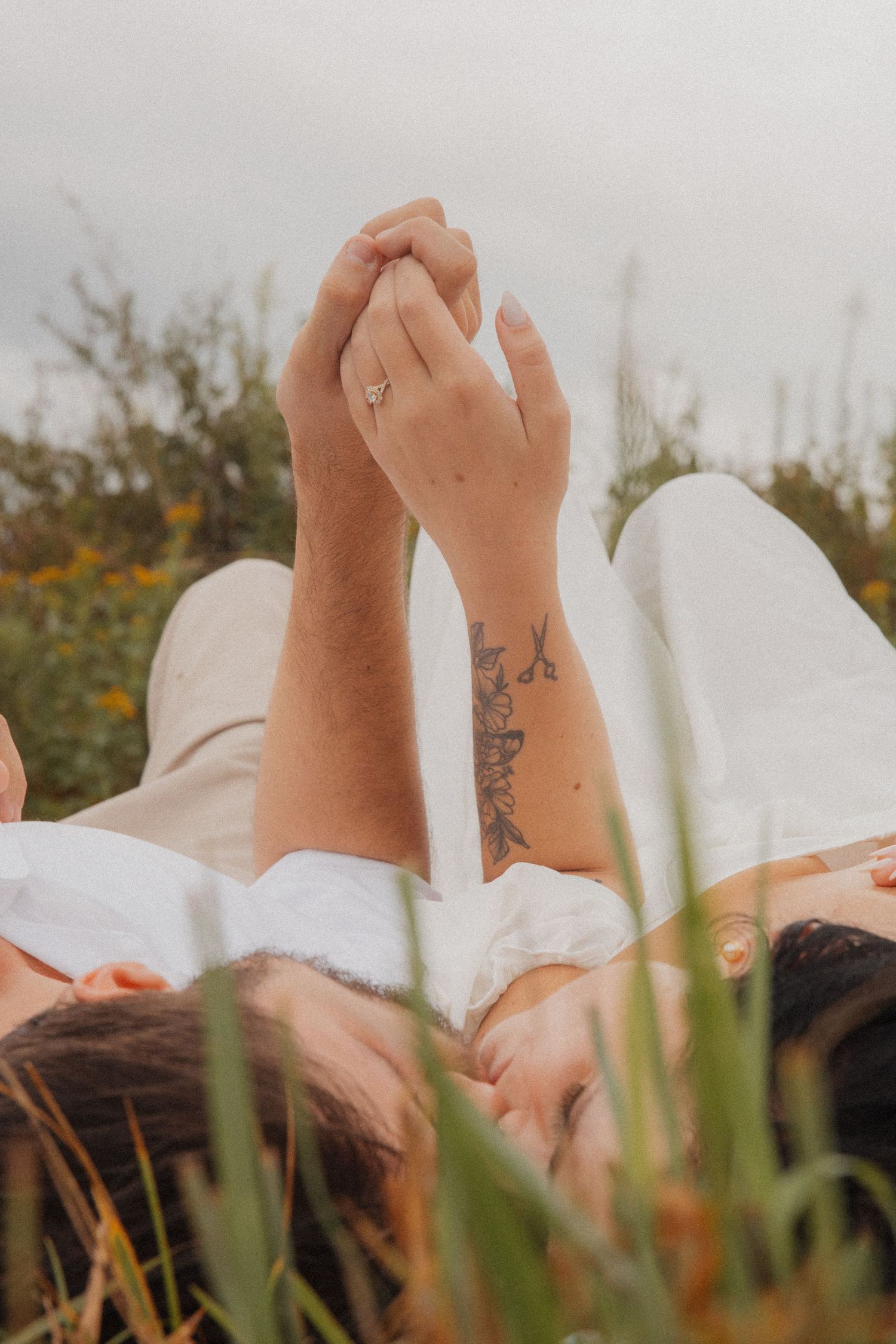 Couple lying in grass holding hands up, man's tattooed arm visible with woman's engagement ring.