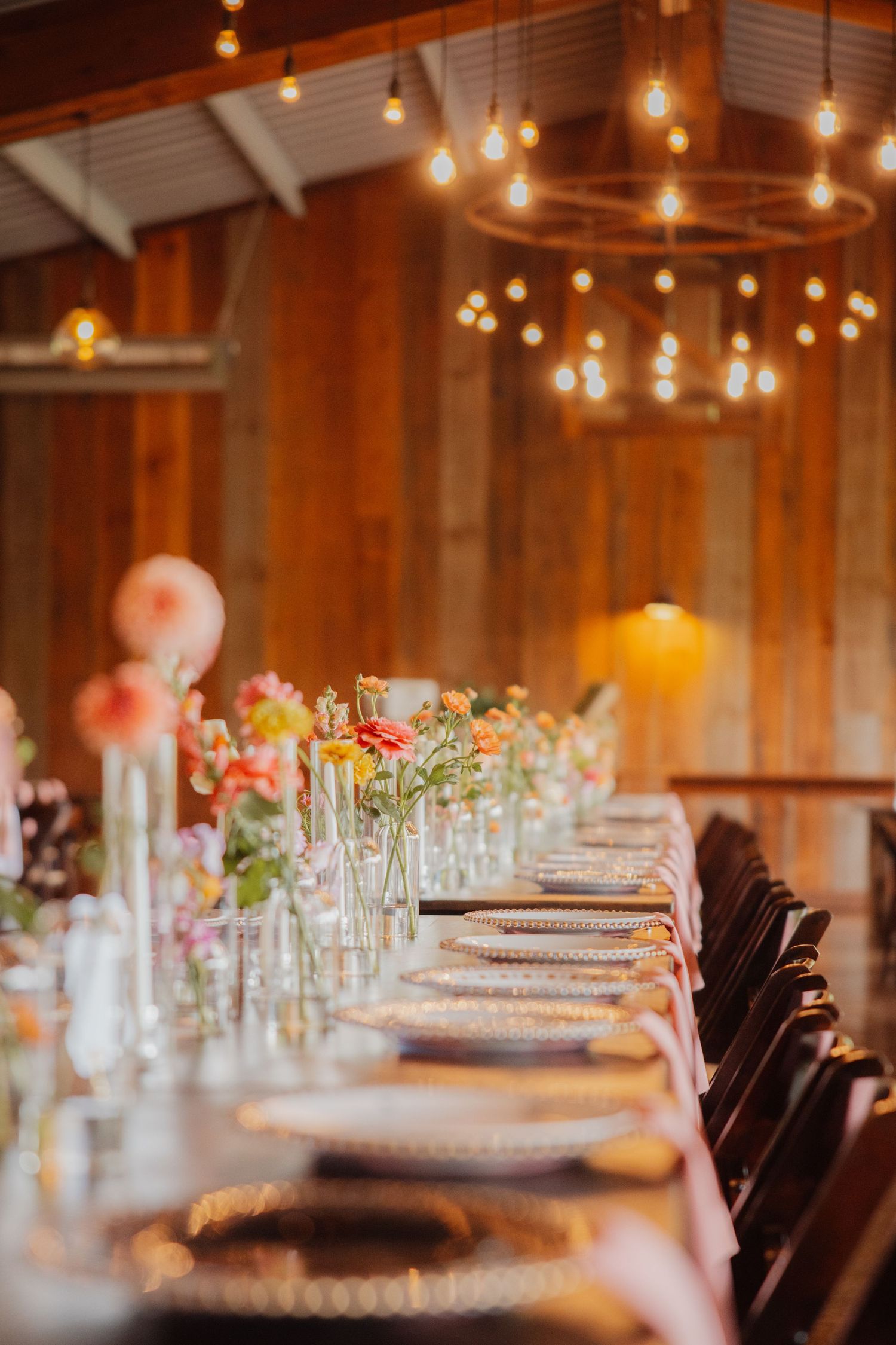 Elegant barn wedding reception table with colorful floral arrangements and warm string lights overhead.