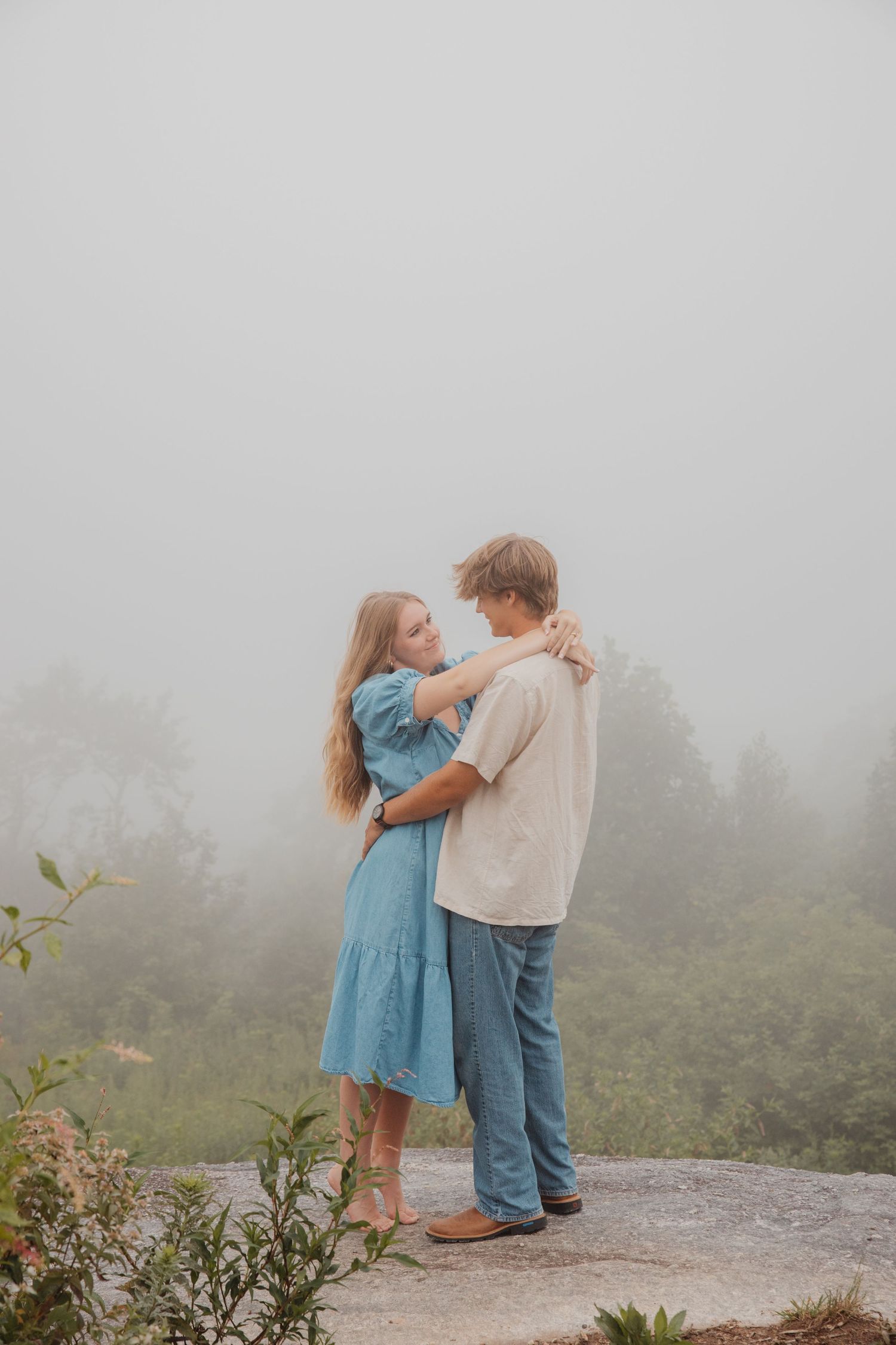 Young couple embracing and gazing at each other on a rocky ledge surrounded by misty green landscape.
