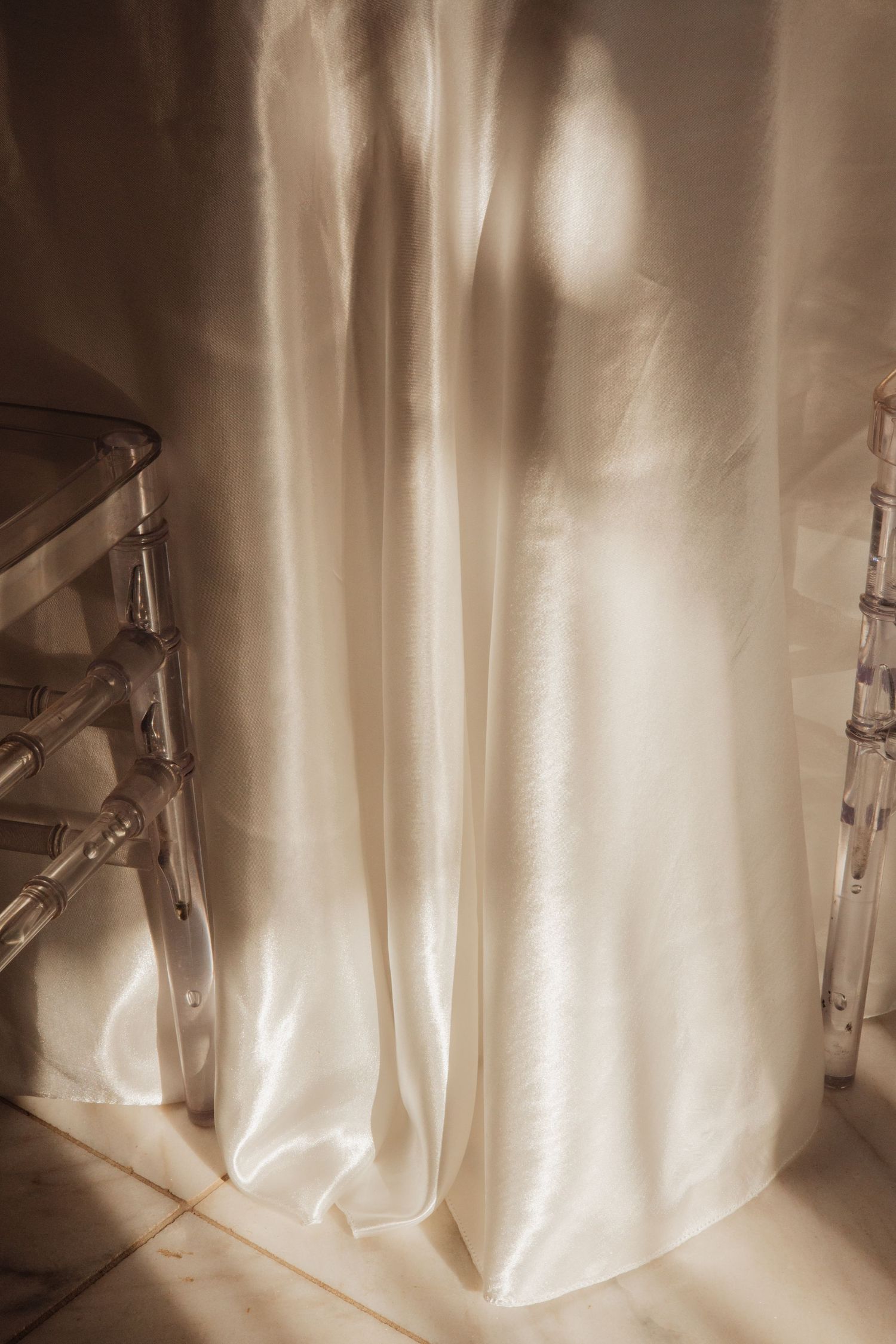 White satin wedding dress draped between two acrylic ghost chairs with warm sunlight casting soft shadows.