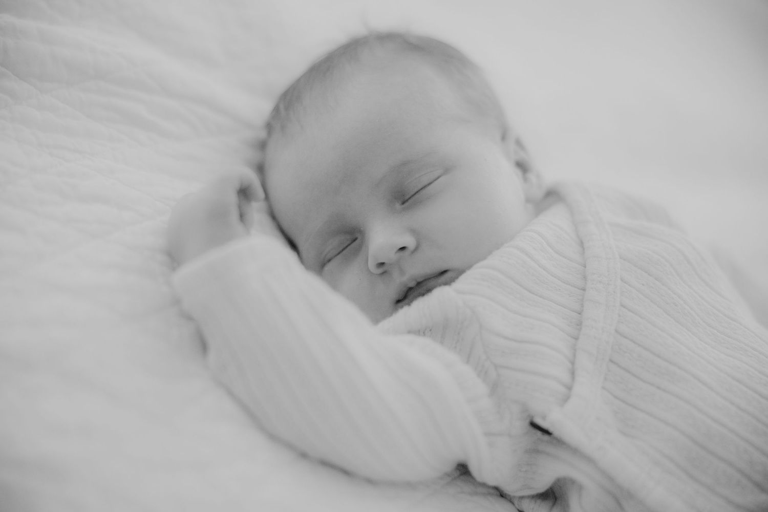 A peaceful newborn baby sleeps wrapped in a soft white swaddle in black and white.