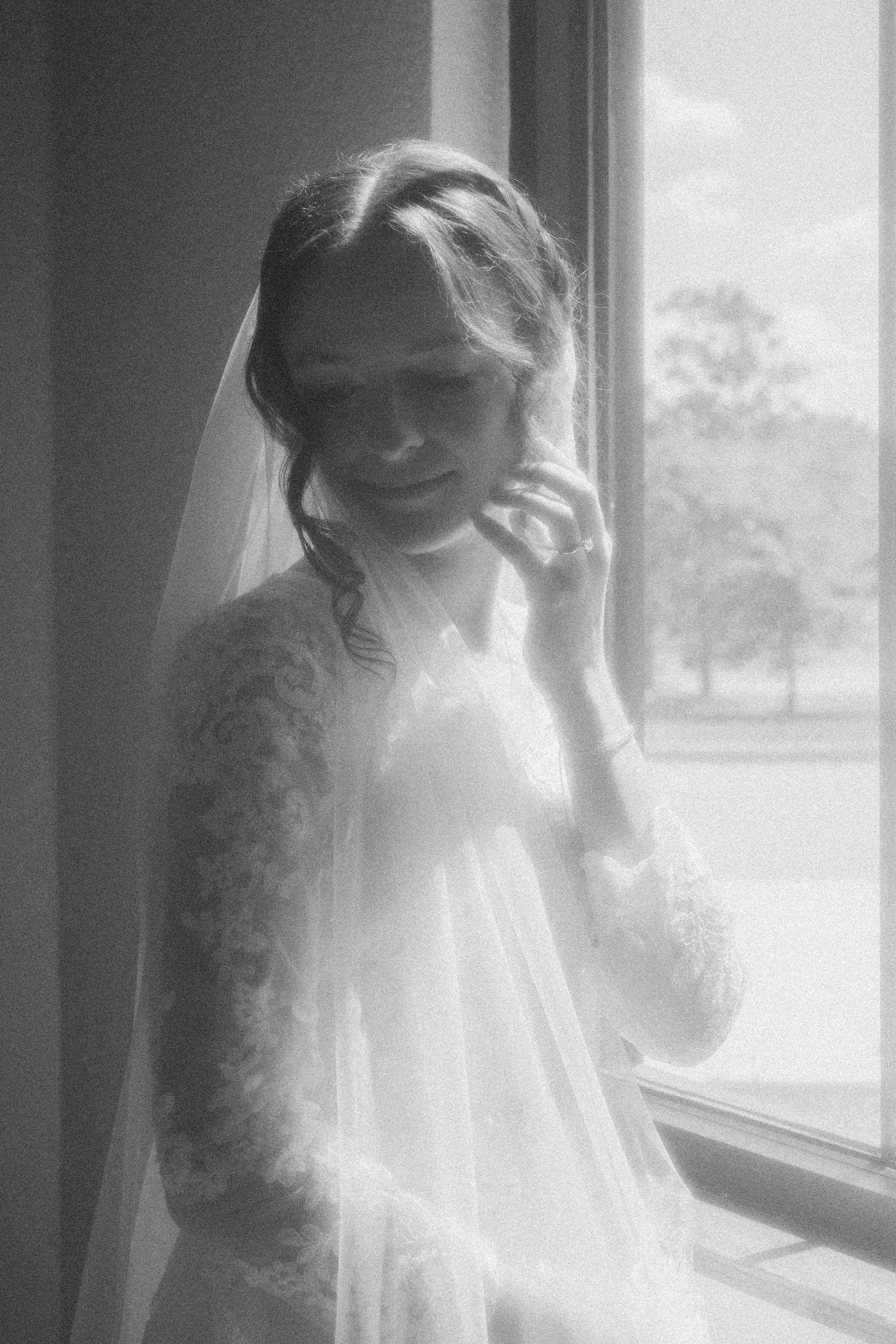 Black and white portrait of a bride in lace wedding dress and veil standing by a bright window.