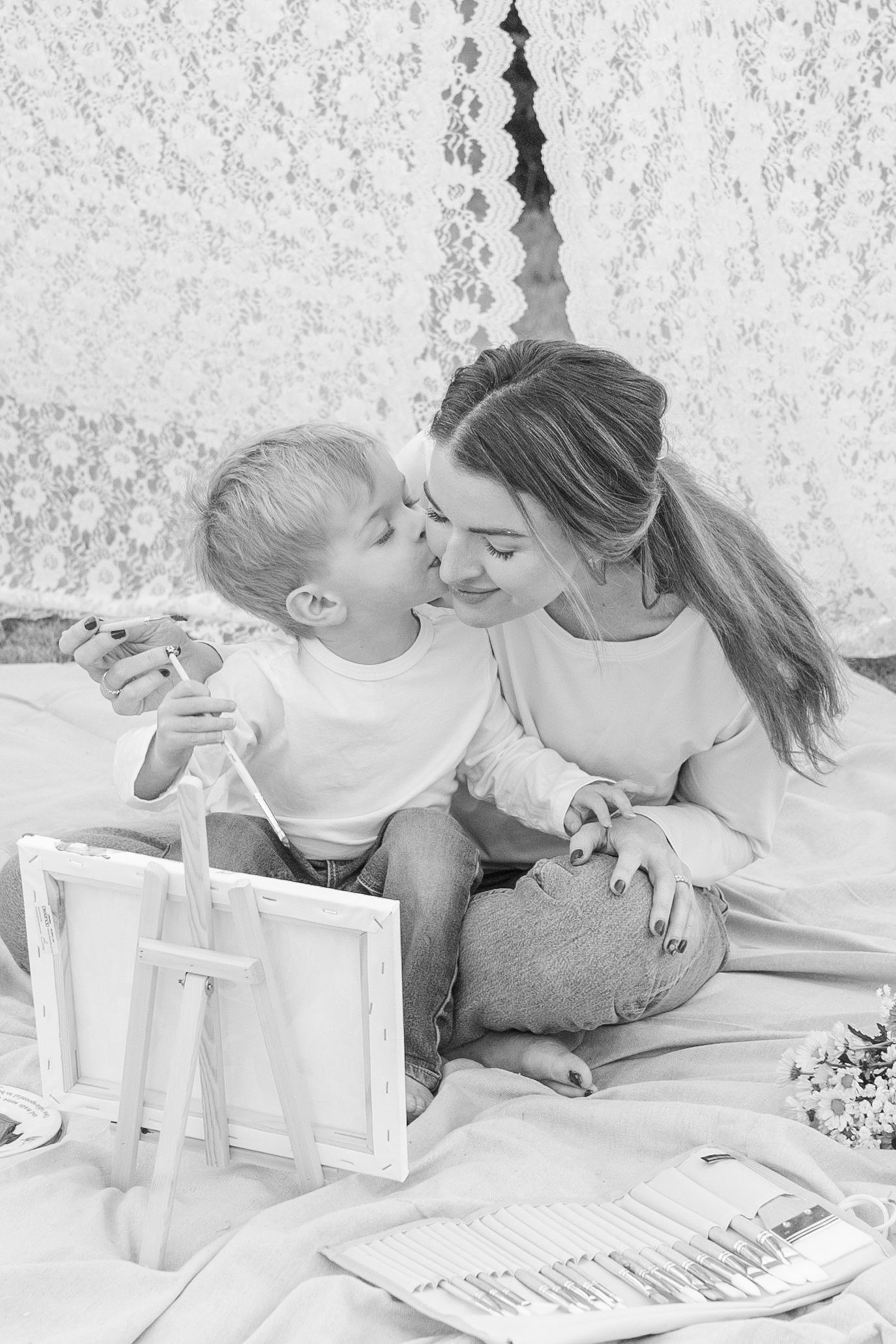 Black and white photo of a young mother and toddler sharing a tender kiss on a bed with picture frames nearby.