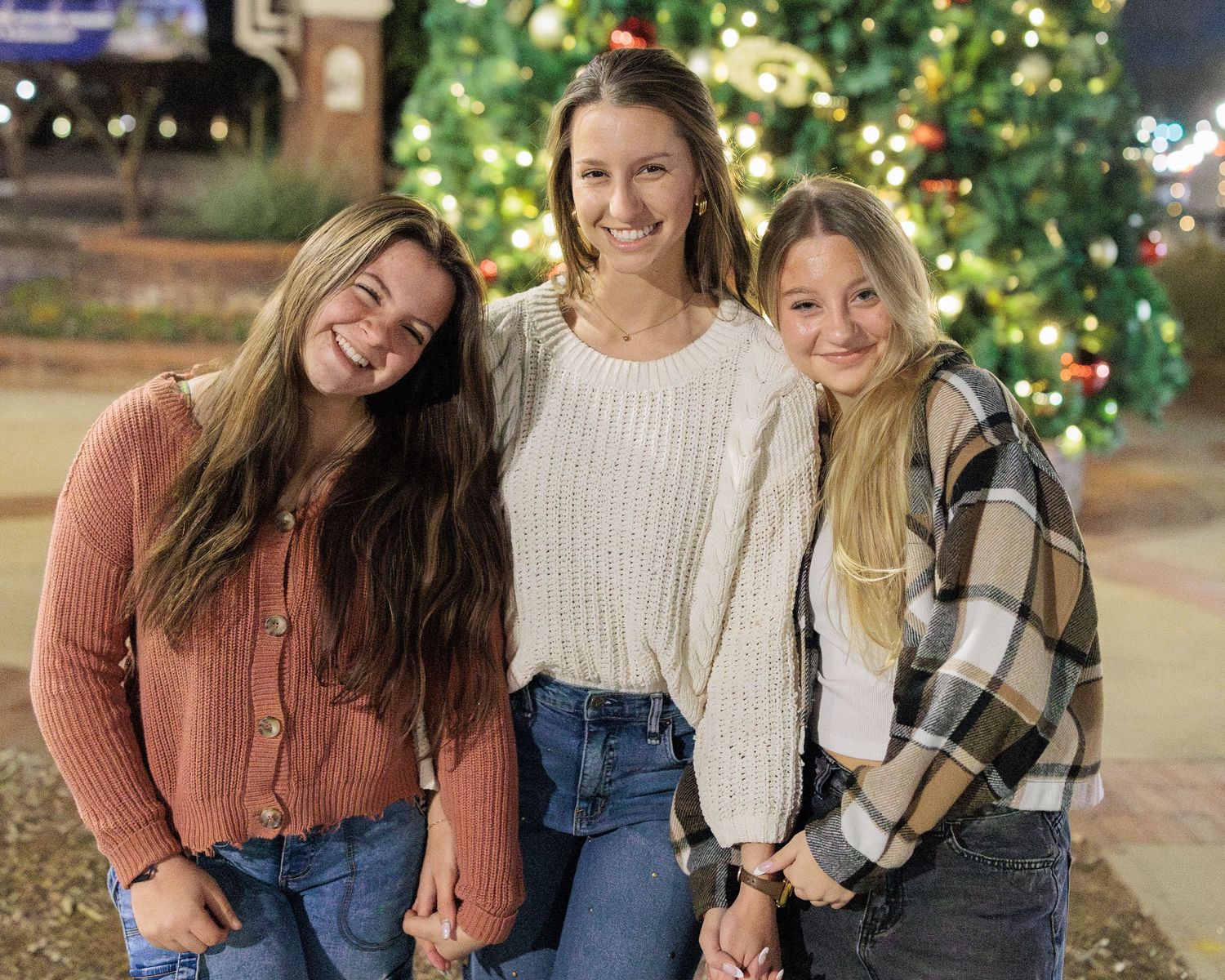 Three smiling young women pose together in front of a decorated Christmas tree outdoors at night.