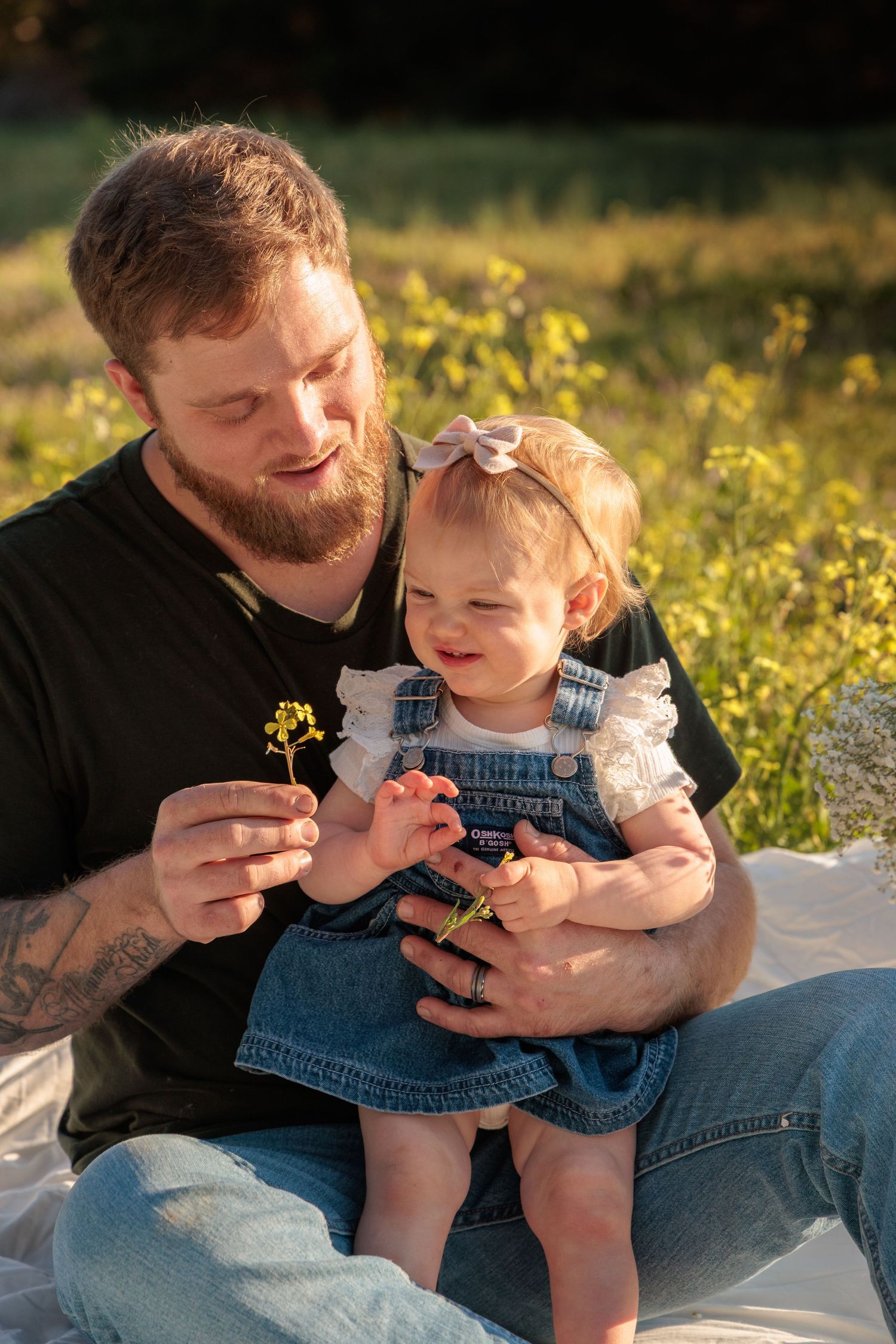 Bearded father sitting outdoors holding baby girl in denim overalls, sharing a yellow wildflower in golden sunlight.