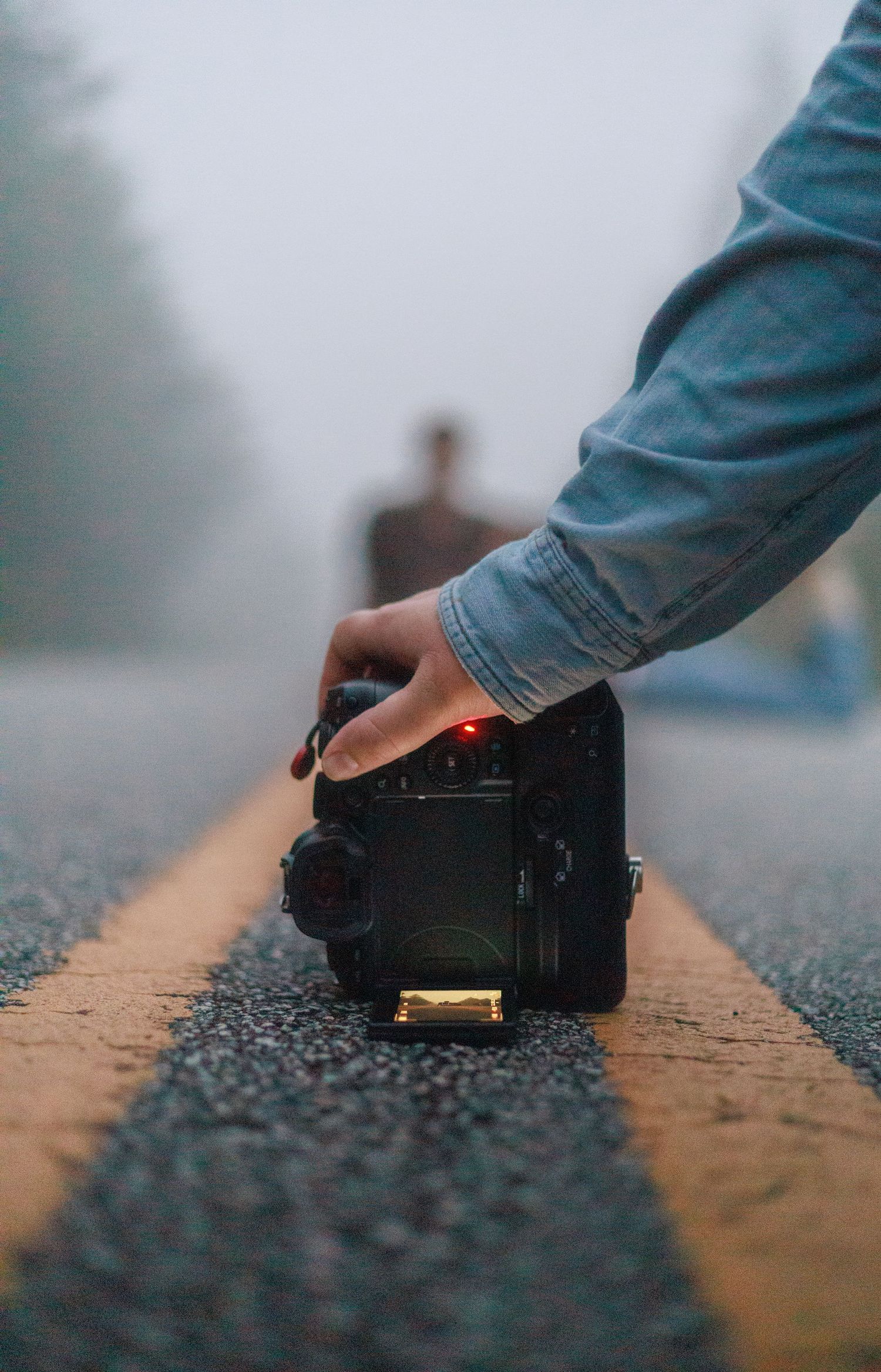 Photographer placing camera on road centerline in foggy conditions with person visible in background.