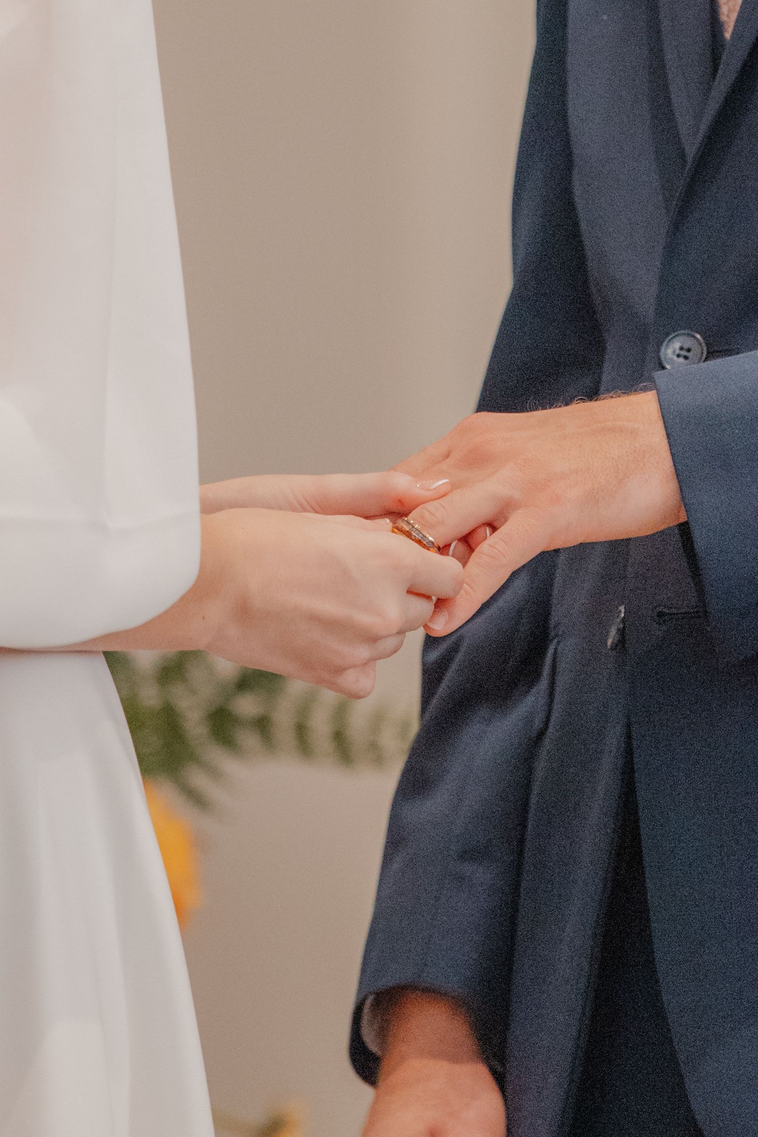 A bride in white places a gold wedding ring on the groom's finger during their wedding ceremony.