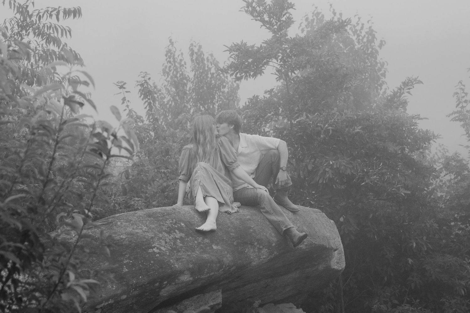 Couple sitting together on a large rock in a foggy forest, sharing an intimate moment.
