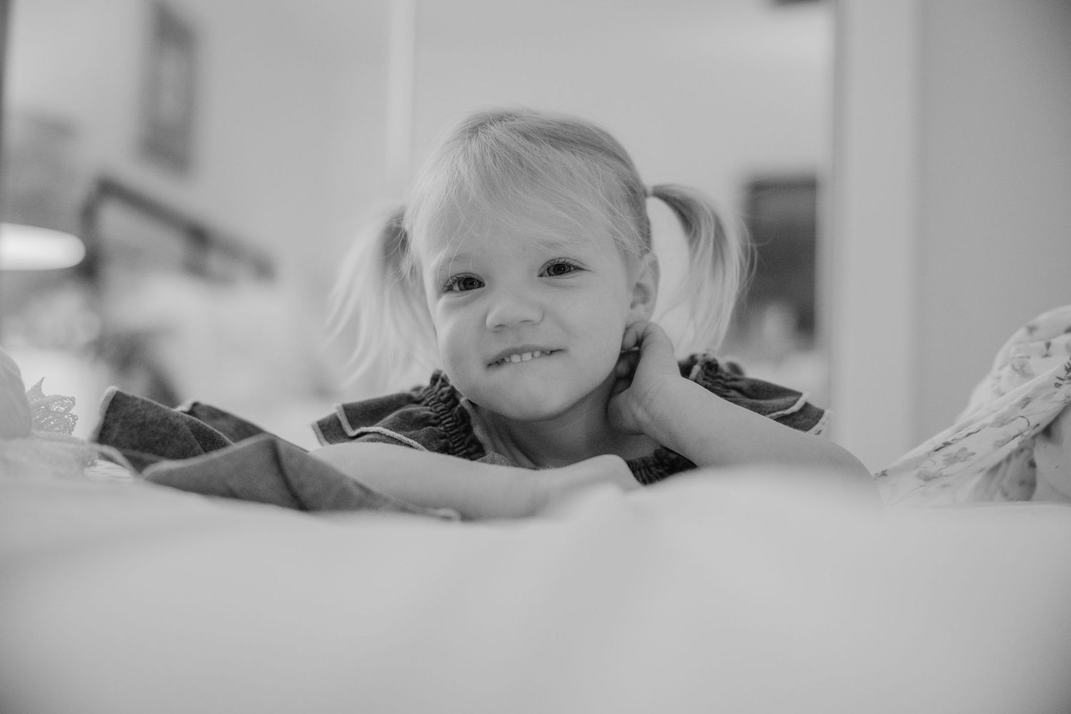 Black and white photo of a smiling toddler girl with pigtails lying on a bed, looking up at the camera.