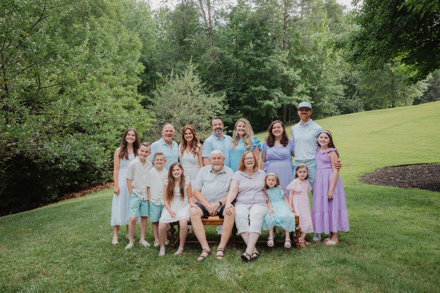 Large family group portrait outdoors on green lawn surrounded by lush trees, wearing coordinated pastel outfits.