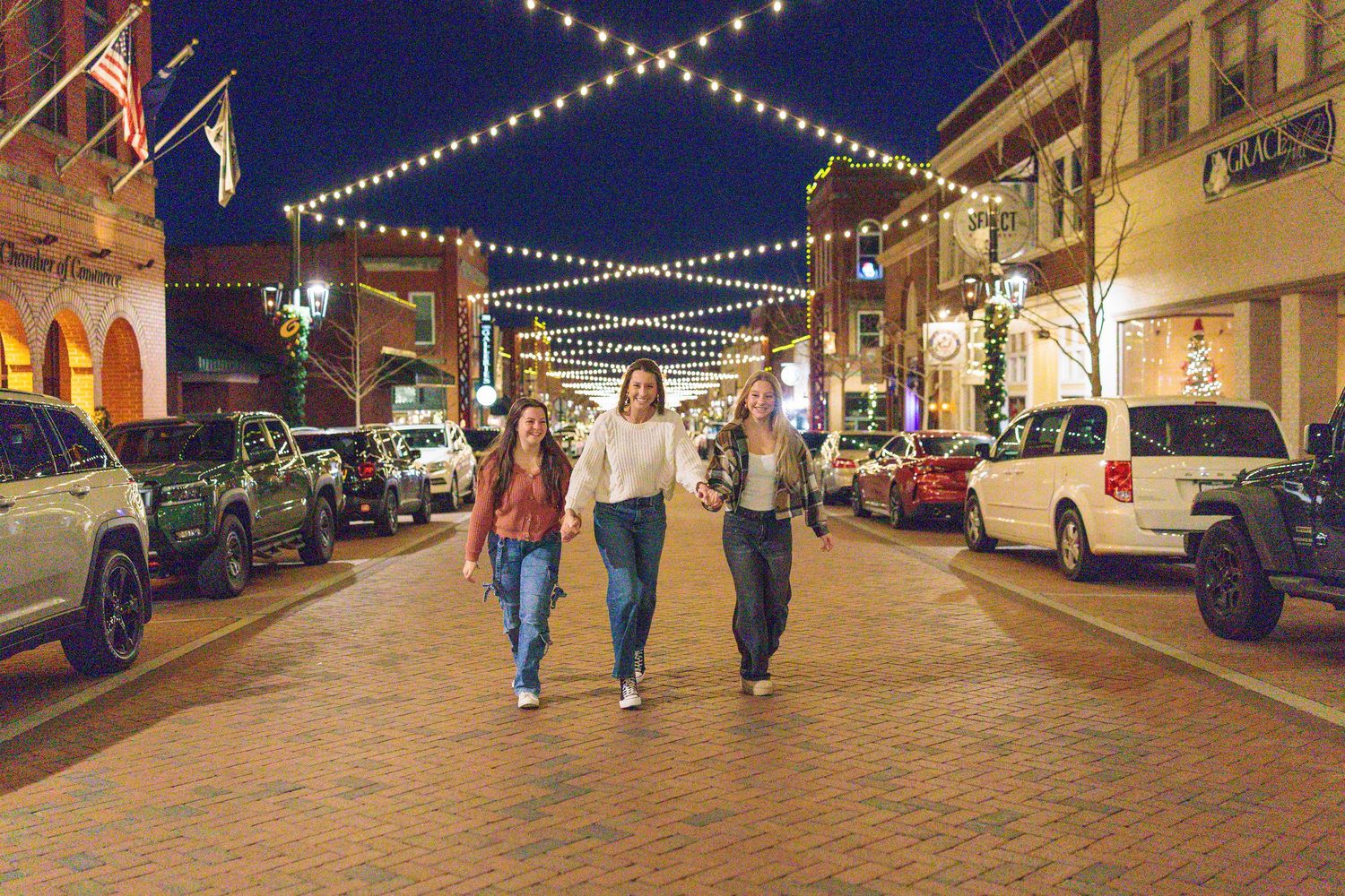 Three women walk together on a charming brick street at night with string lights overhead in a vibrant downtown district.