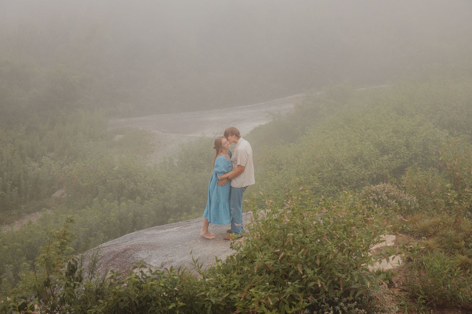 Young couple sharing a romantic kiss while embracing on a rocky overlook surrounded by misty greenery.