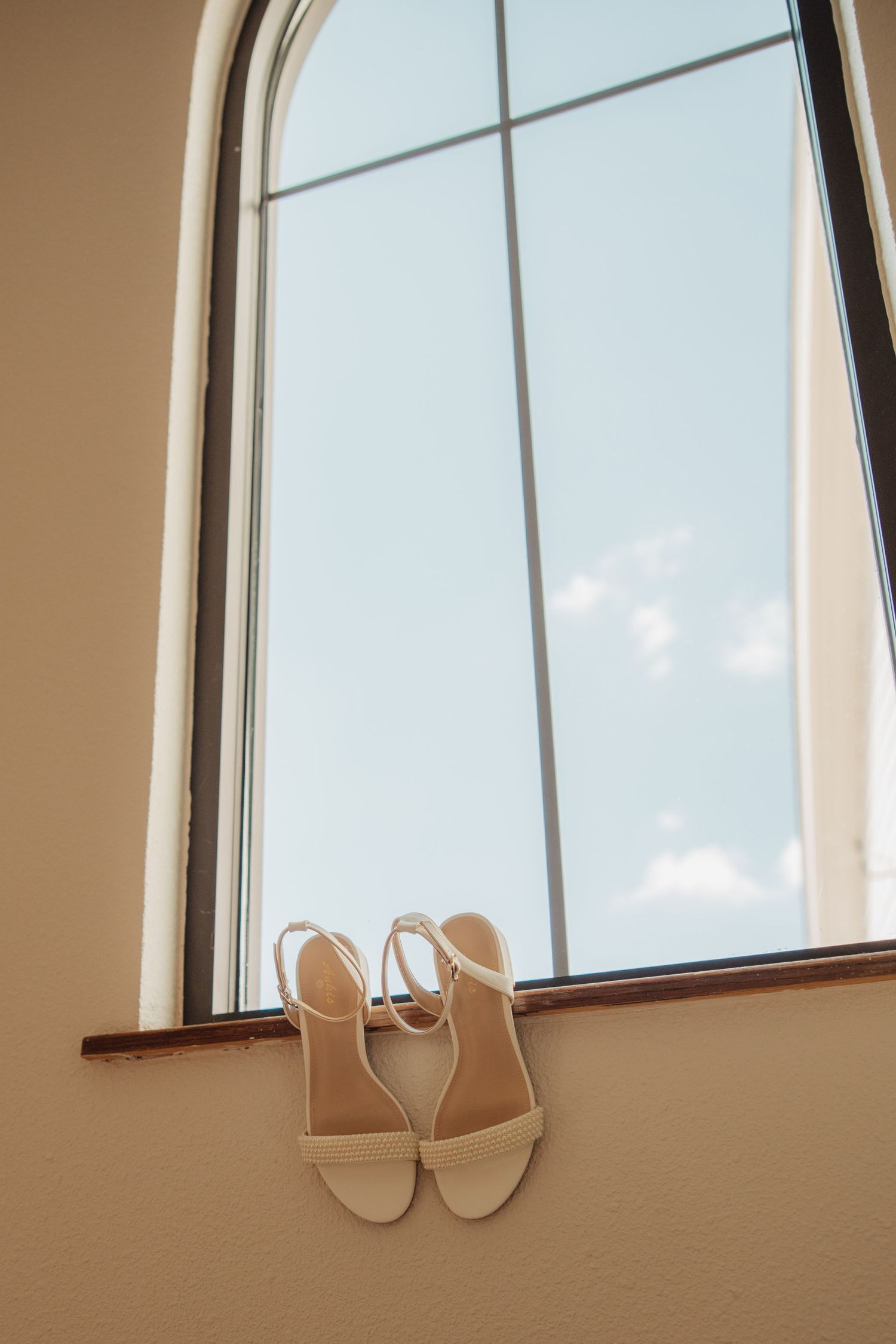 Elegant nude strappy bridal heels displayed on a window ledge with arched window and blue sky backdrop.