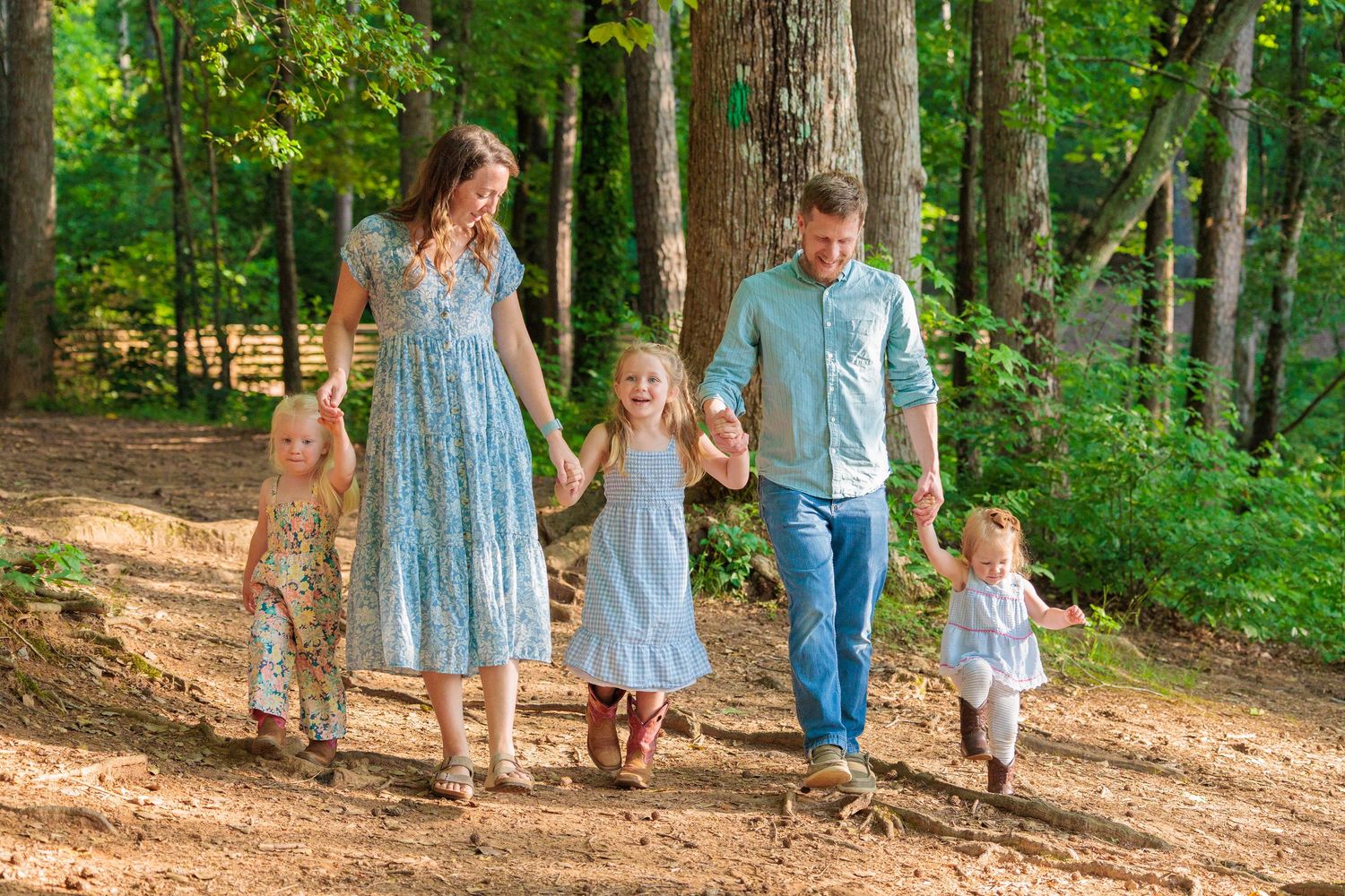 Family of five walking hand-in-hand on a sunny forest path, parents and three young children in coordinating outfits.