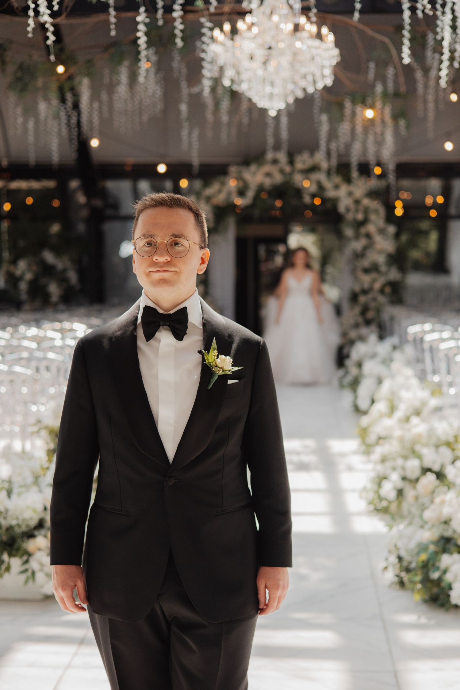 Groom in black tuxedo with bow tie stands at altar as bride approaches down floral aisle.