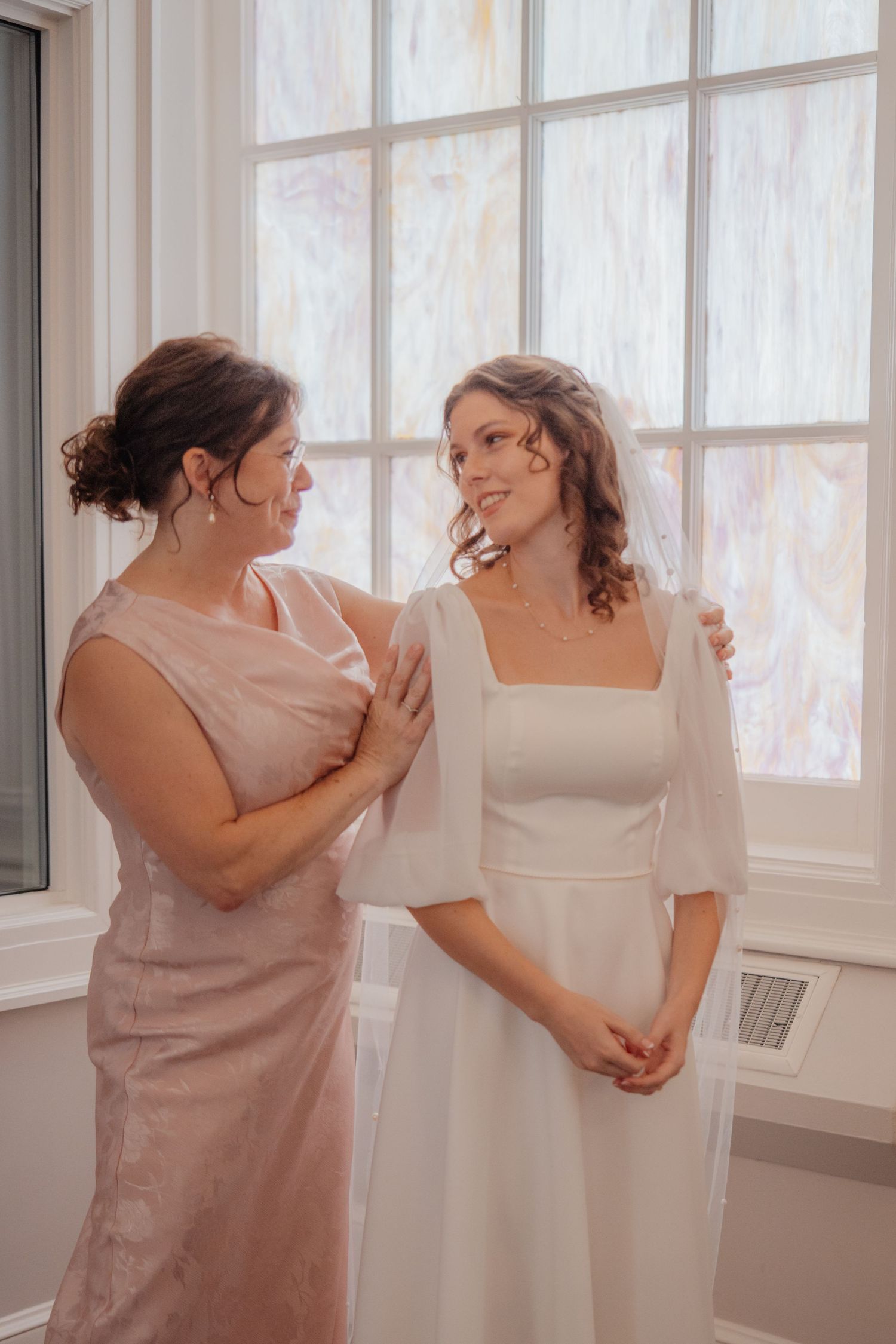 A bride in a white dress with flutter sleeves shares a tender moment with a woman in a pink dress by a window.