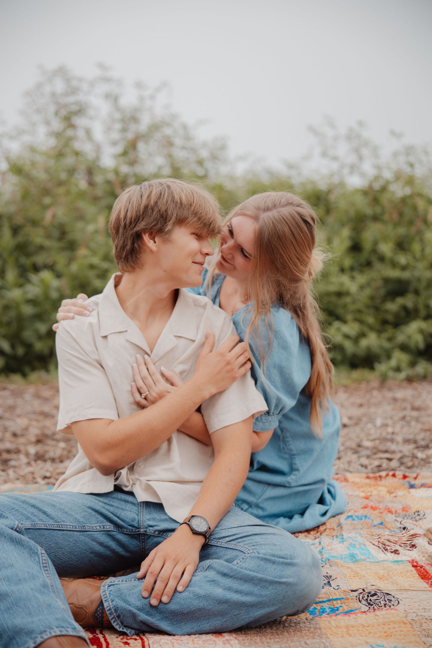 Young couple sitting on colorful blanket outdoors embracing and smiling at each other in misty garden setting.