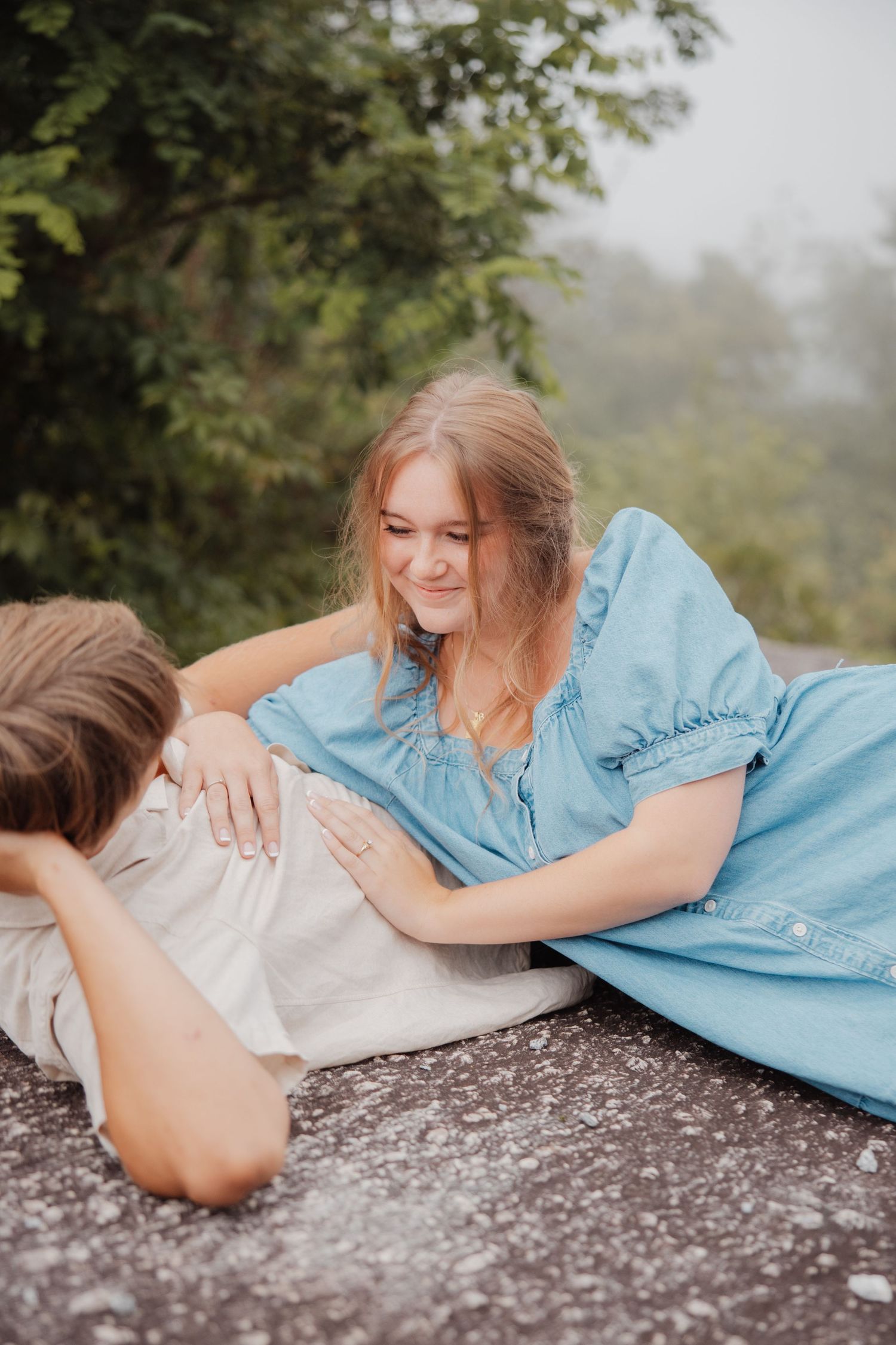 Young woman in blue dress smiling at man lying in her lap outdoors on rocky surface with green trees behind.