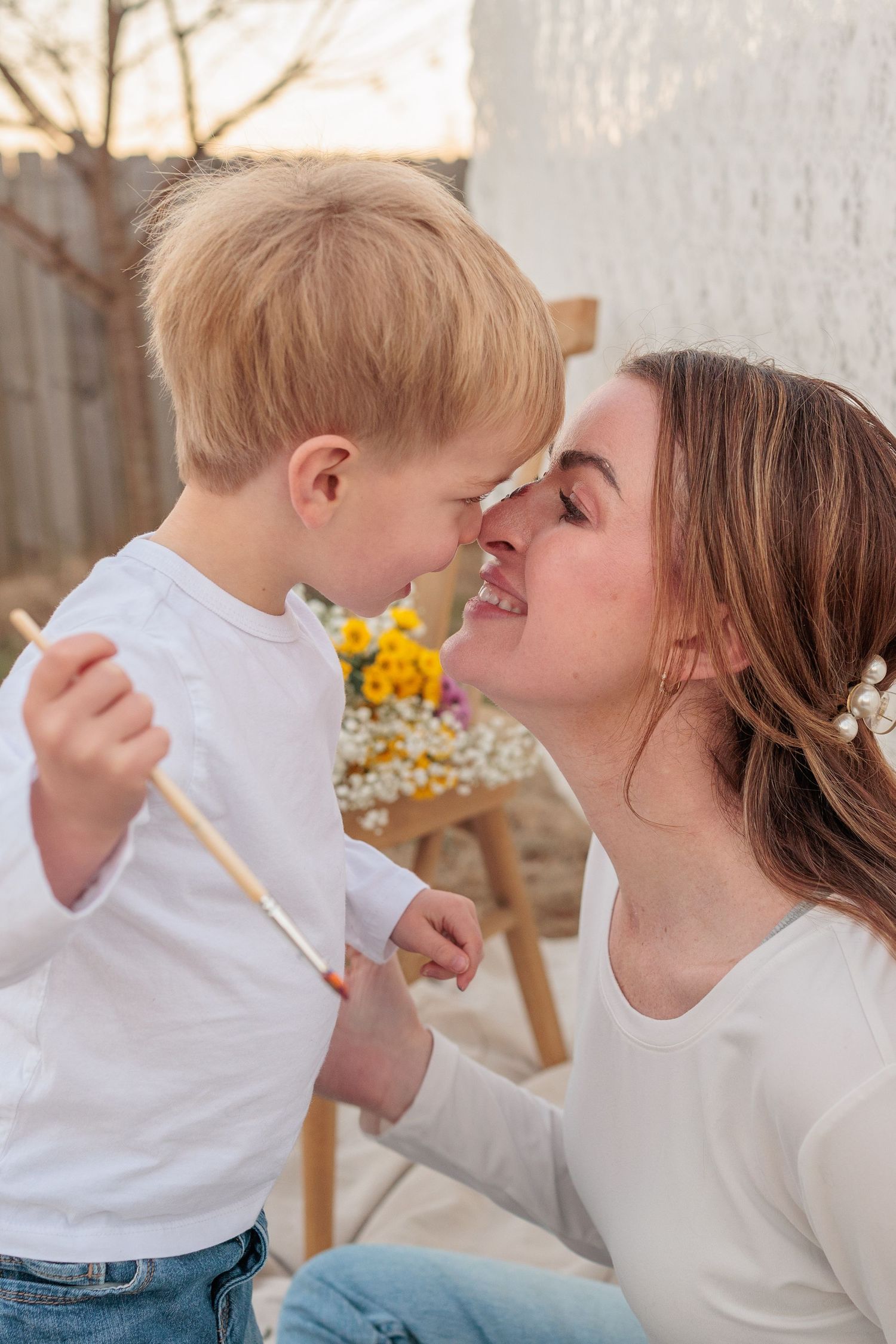 A mother and young blonde boy touch noses and smile outdoors, with yellow flowers in the background.