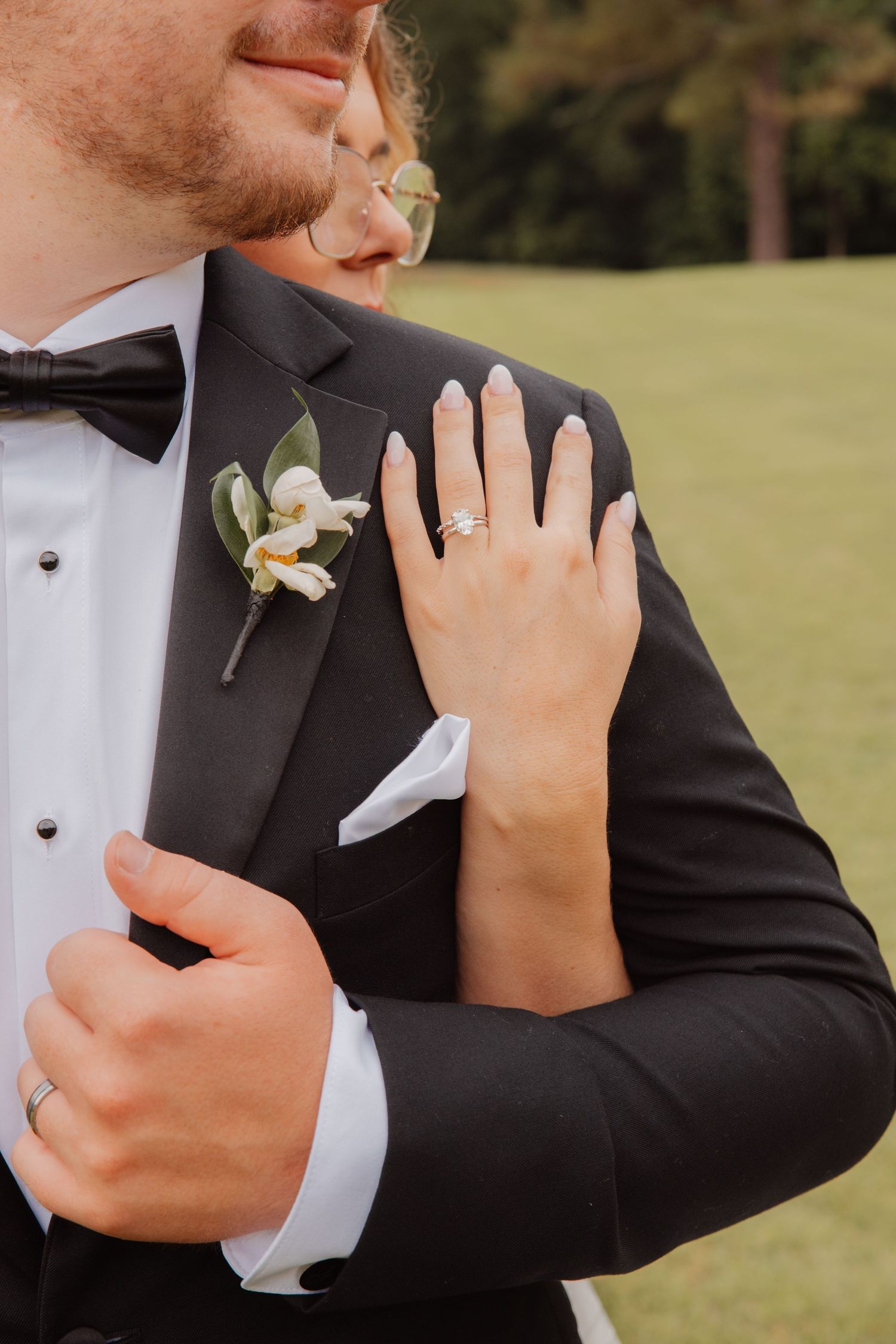 Close-up of bride showing diamond engagement ring while embracing groom in black tuxedo with floral boutonniere.