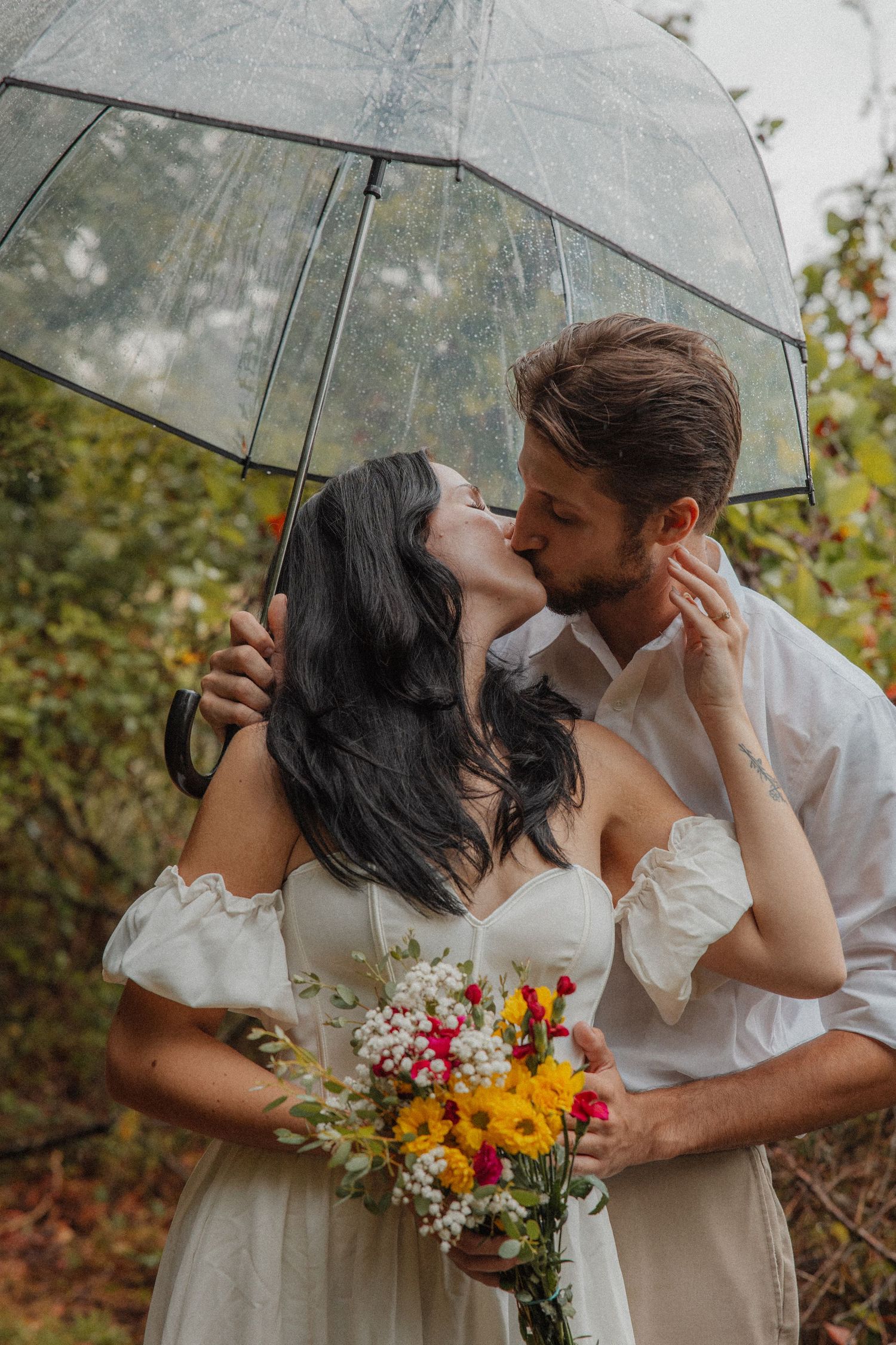 Couple sharing a kiss under a clear umbrella, woman holding a colorful wildflower bouquet.