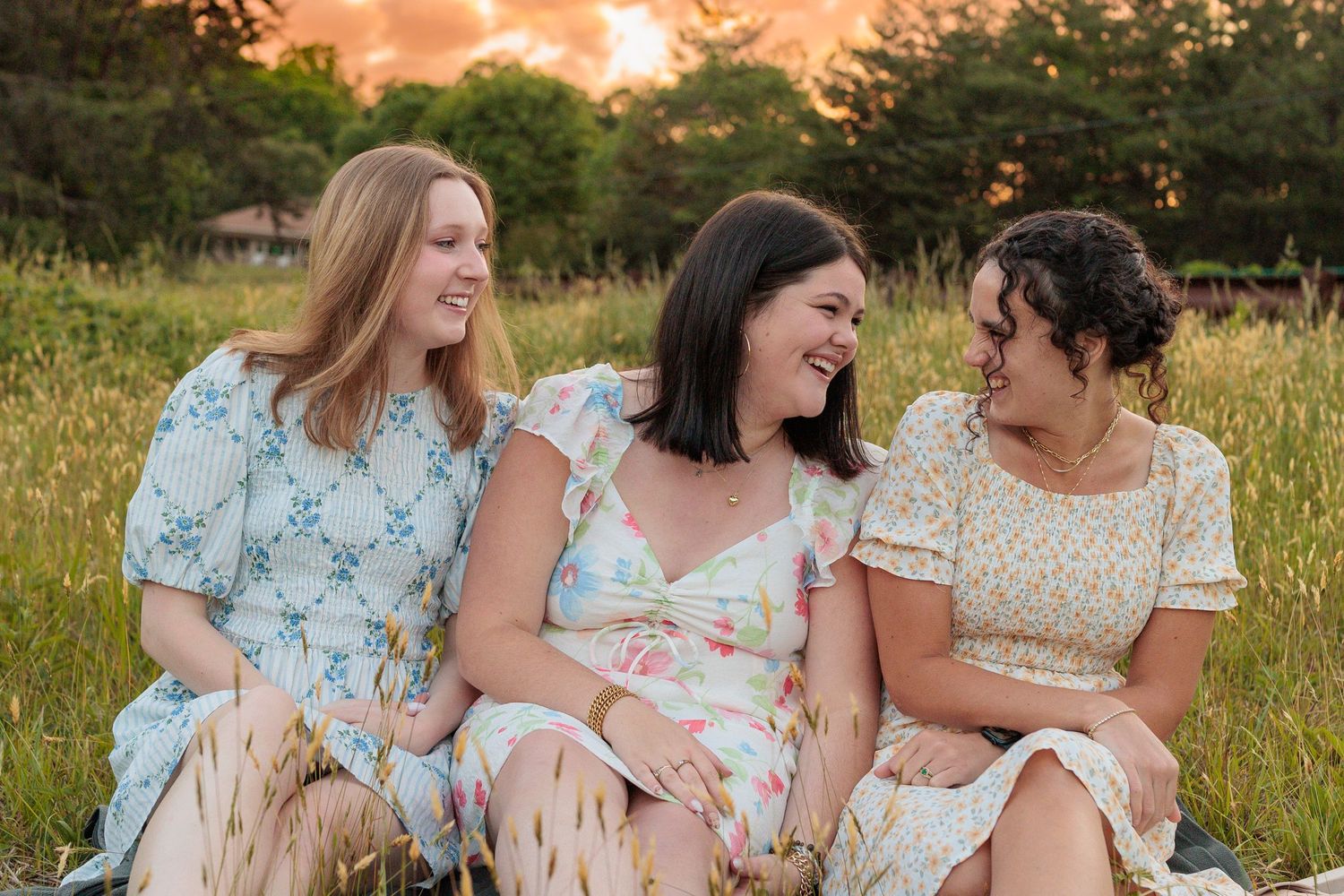 Three women in floral dresses laugh together while sitting in a golden field at sunset.