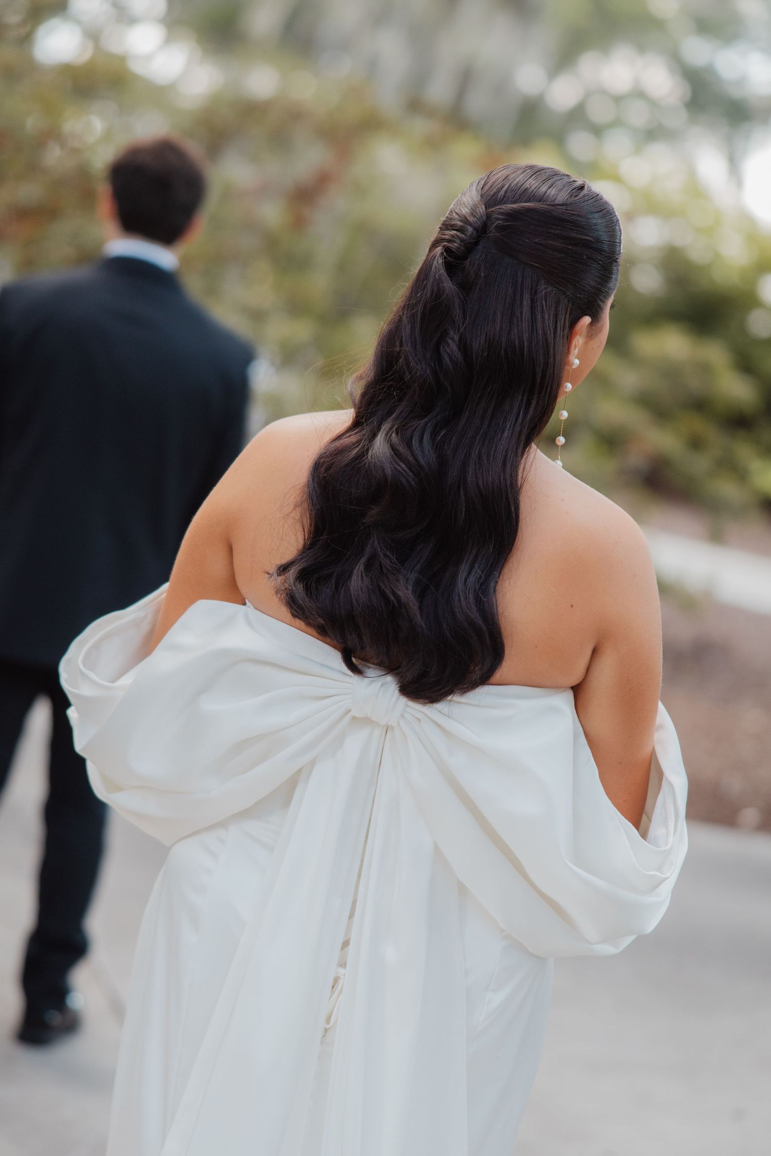 Bride in elegant strapless white gown with large bow detail, dark wavy hair, facing away.