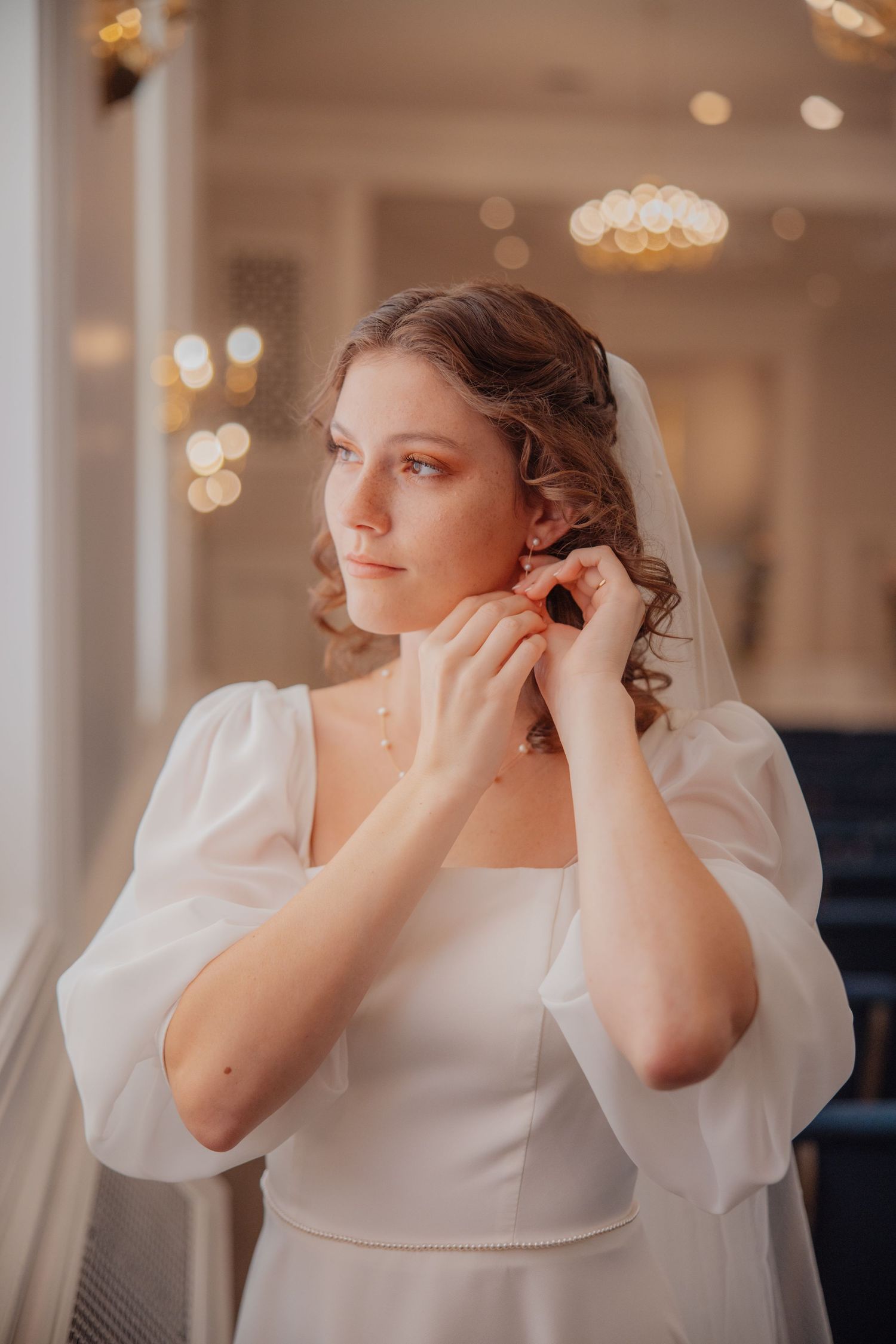 A bride in a white square-neck gown with puff sleeves adjusts her earring while wearing a veil.