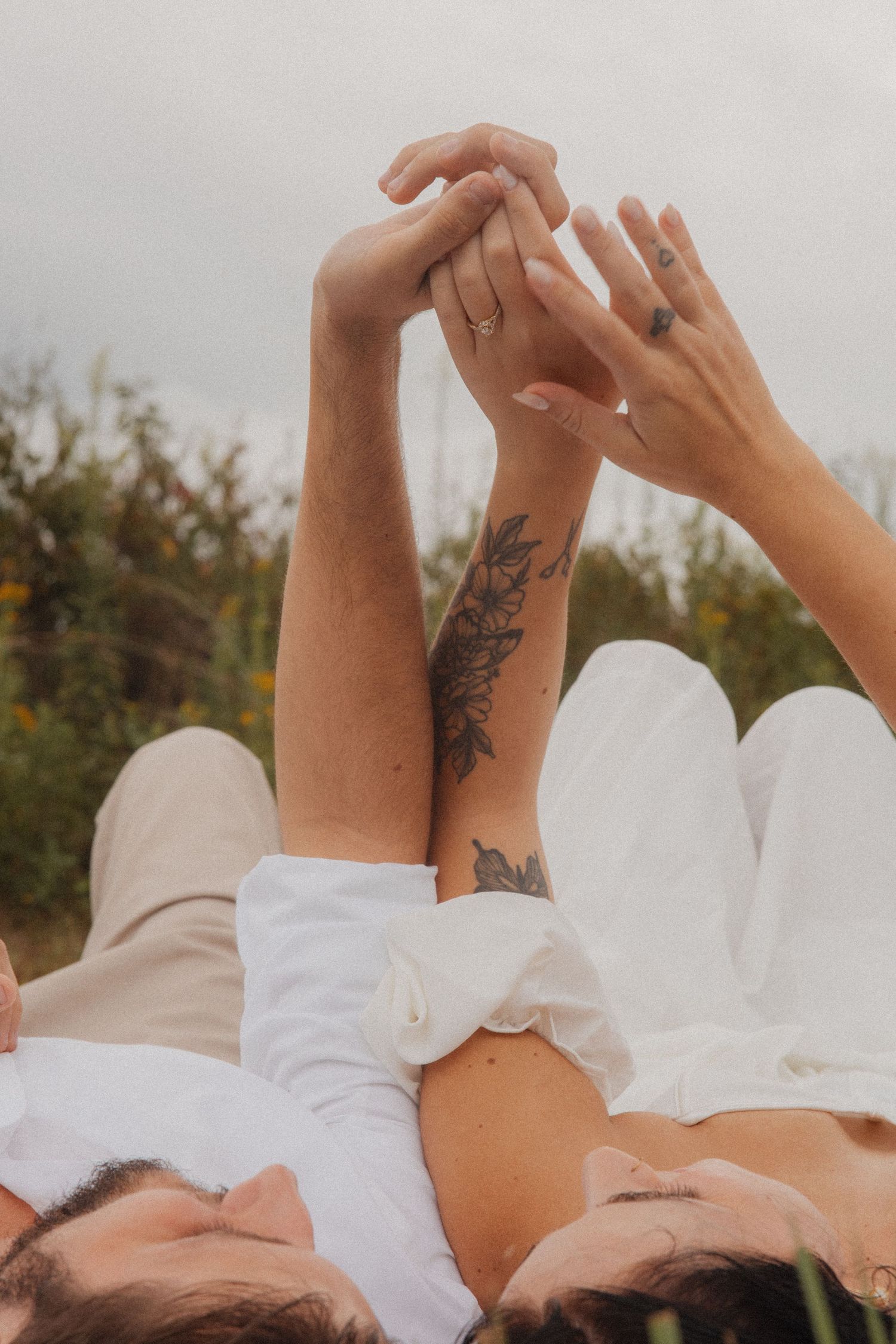 Couple lying in a field holding hands up, showing tattoos and wedding rings against cloudy sky.
