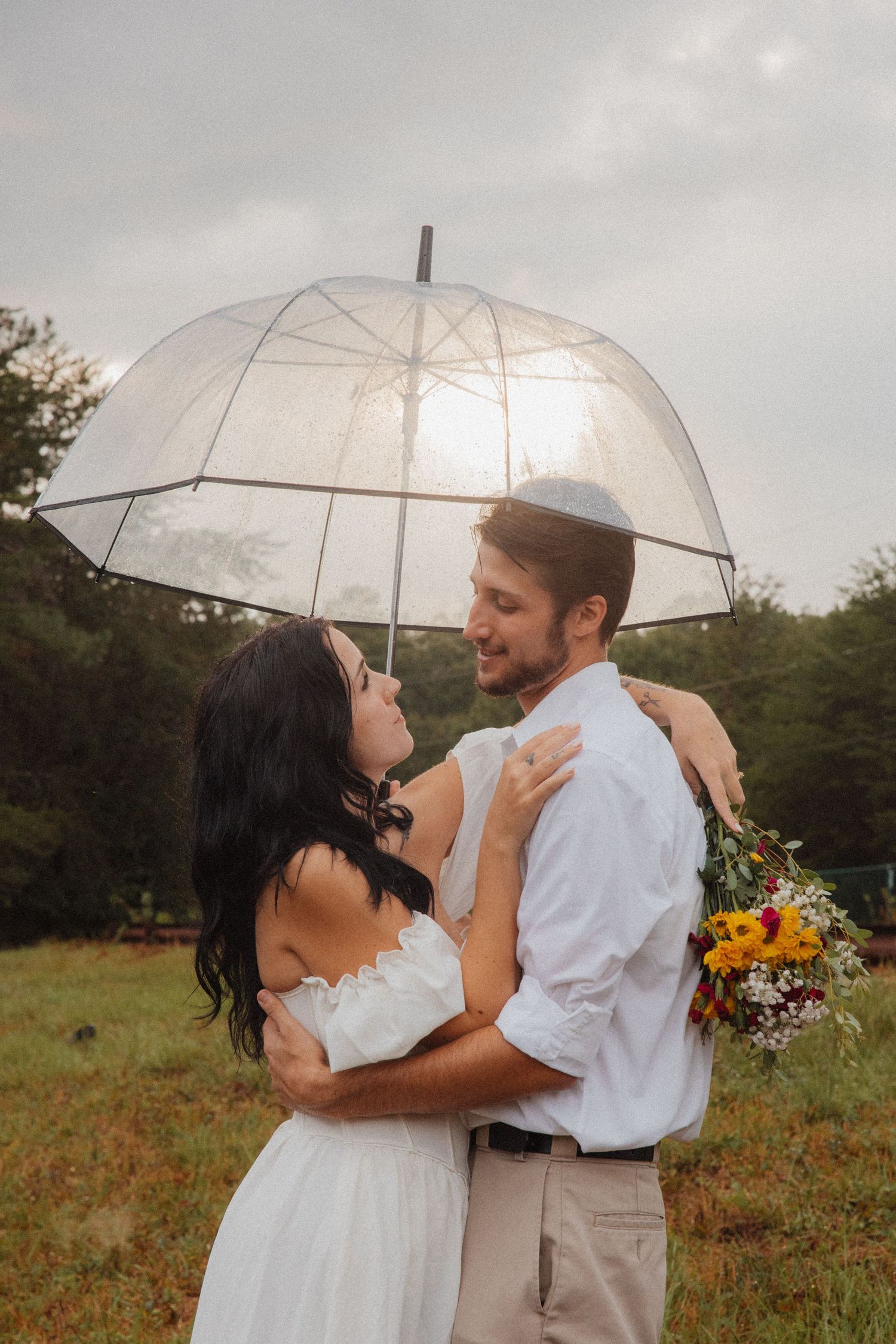 Smiling couple embracing under clear bubble umbrella holding wildflower bouquet in rainy field.