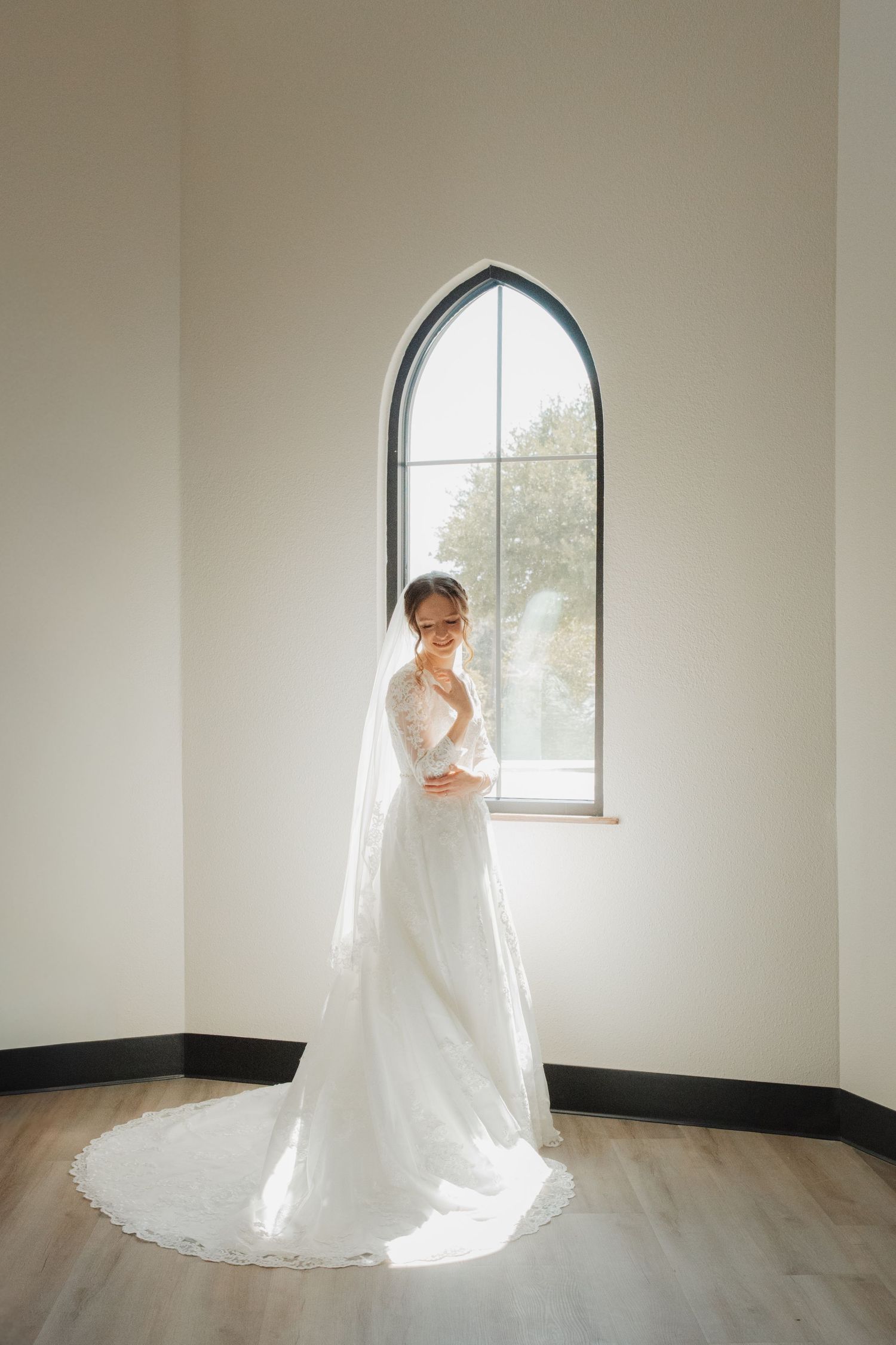 Bride in long-sleeve lace wedding gown with cathedral veil standing in sunlit chapel room with arched window.