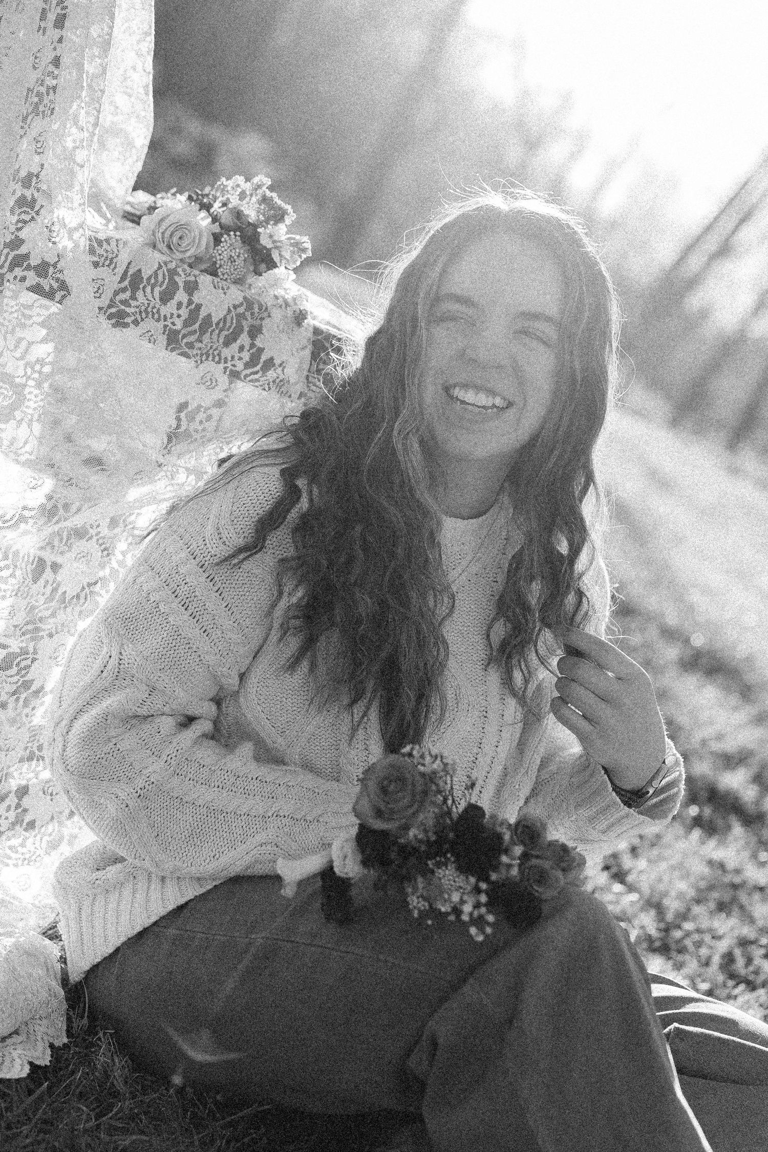 Young woman with long curly hair smiling joyfully outdoors holding flowers in black and white photo.