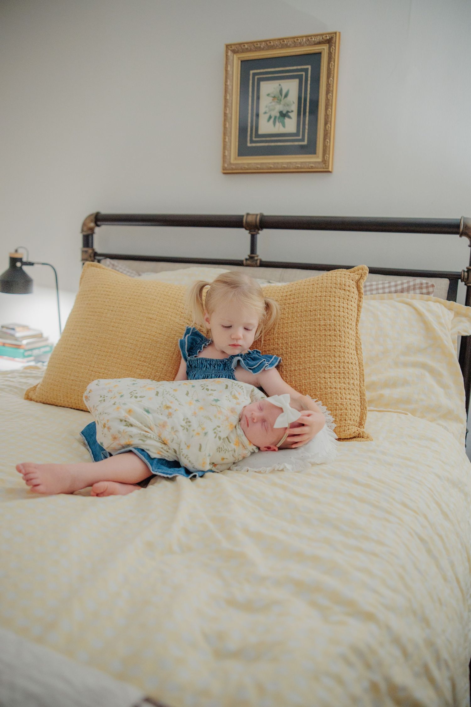 Toddler girl in denim dress holding newborn baby sibling on yellow bedding with gold pillows.