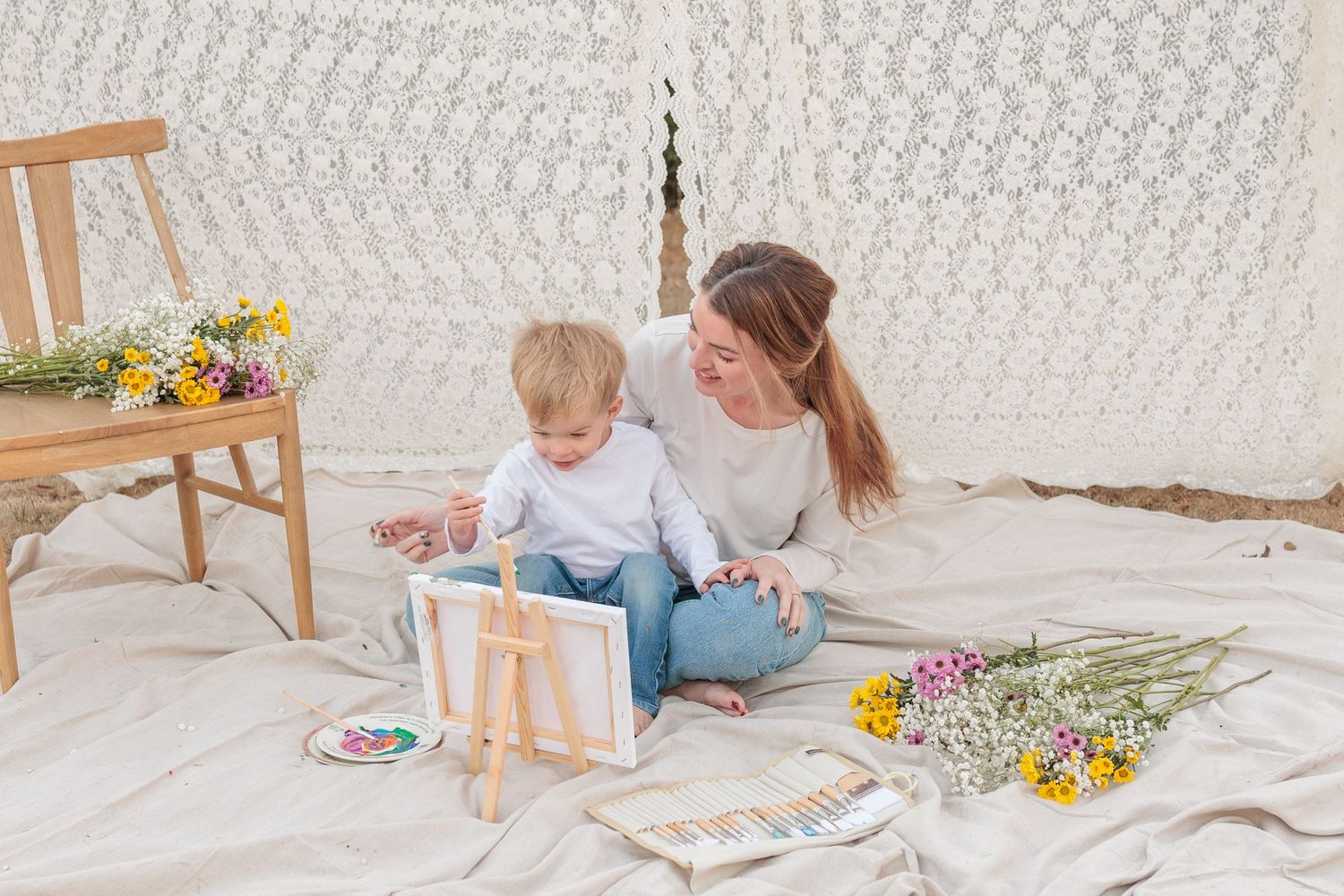 Mother and toddler sitting on blanket painting together surrounded by wildflowers in a cozy boho tent setting.